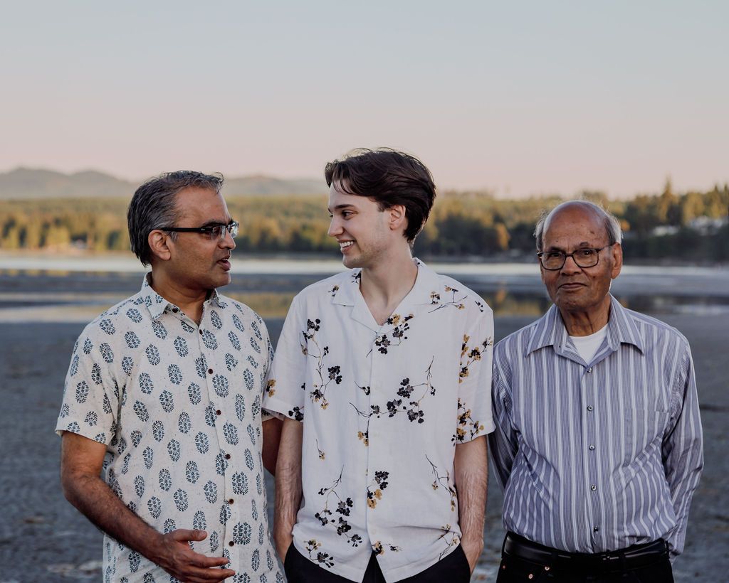 Three men stand together outdoors near water at golden hour, middle man in floral shirt smiling at left.