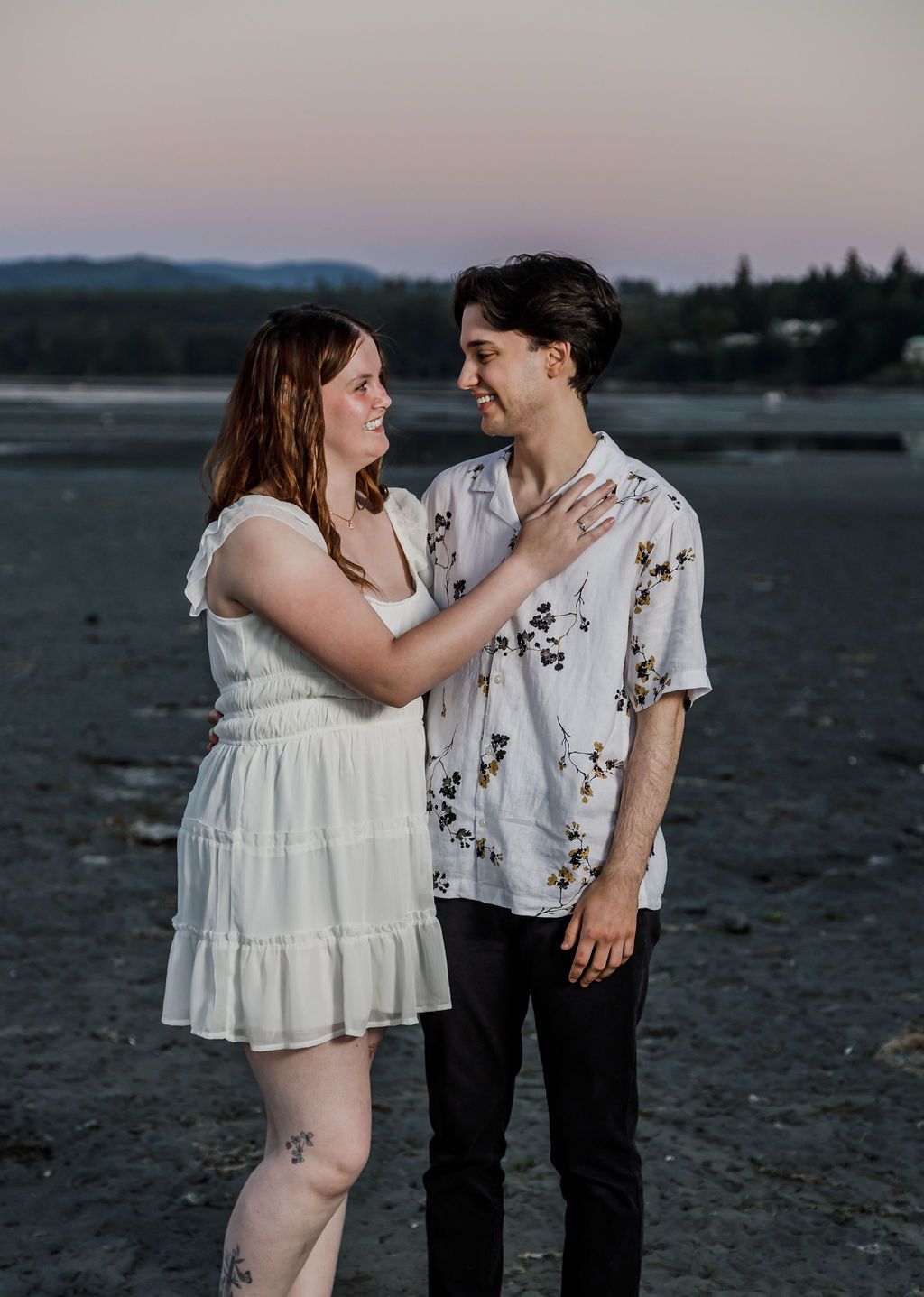 Couple sharing a romantic moment on a beach at dusk, woman in white dress touching mans chest.