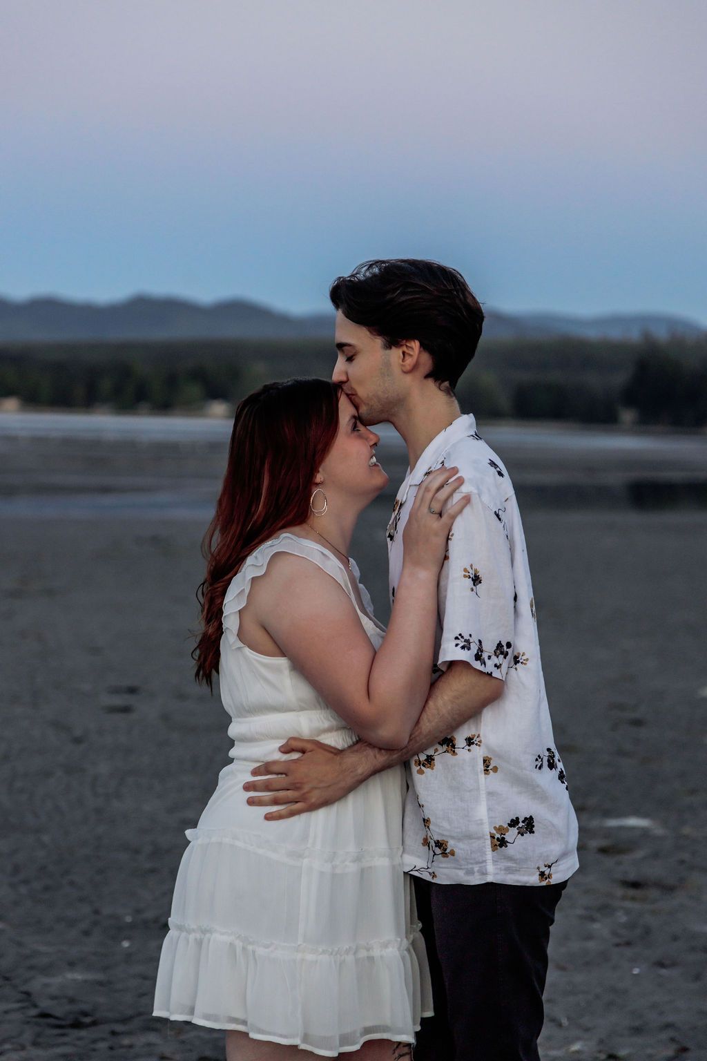 Romantic couple about to kiss on a beach at twilight with mountain silhouettes behind them.