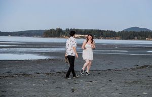 Couple walking together on a sandy beach during daytime with forested hills in the background.