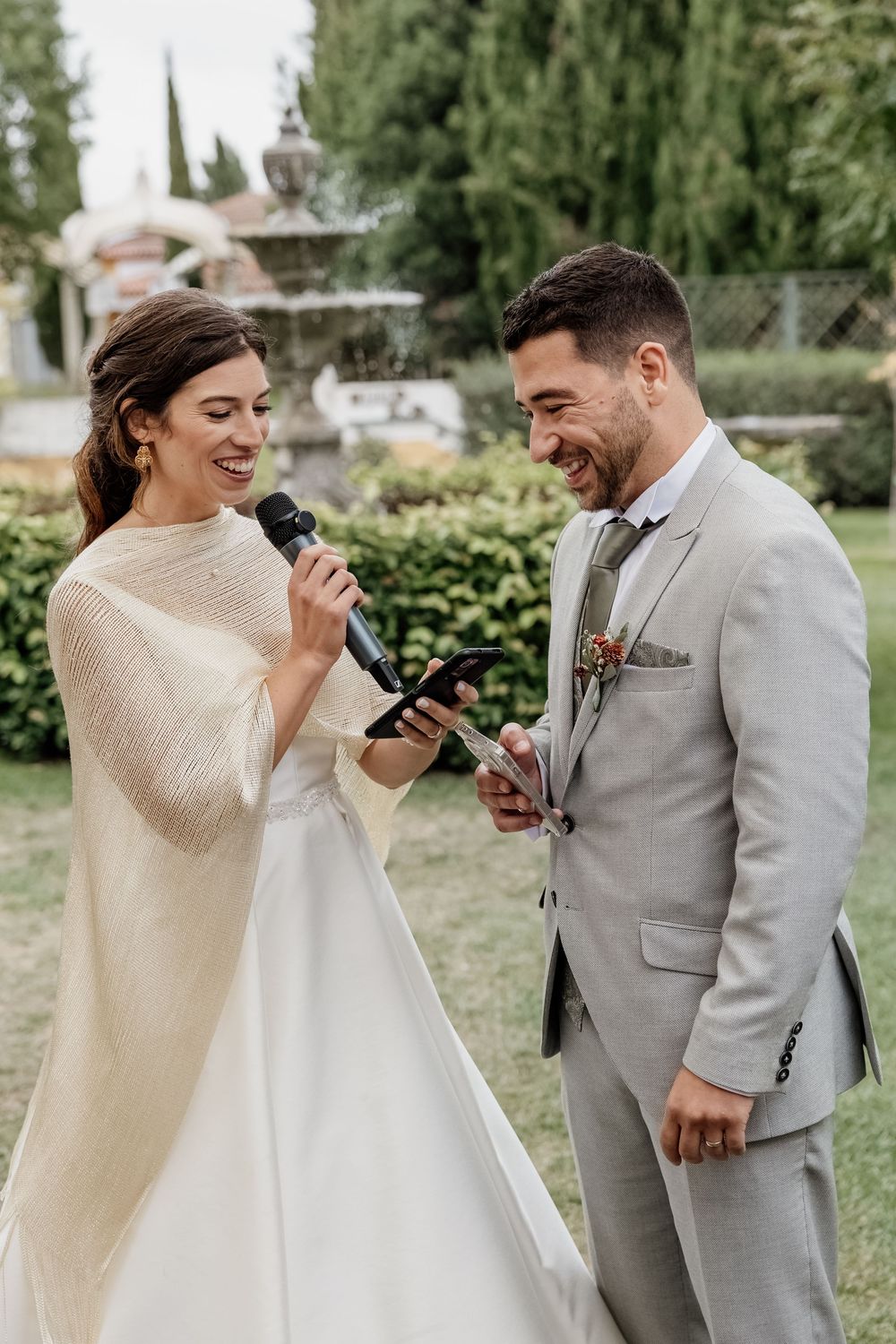 Bride reads wedding vows into microphone while groom smiles during outdoor garden ceremony.