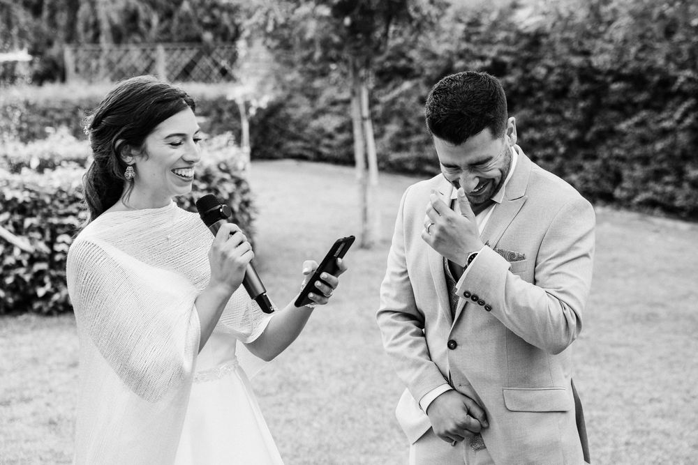 Black and white wedding photo of bride reading vows from phone while emotional groom wipes tears during outdoor ceremony.