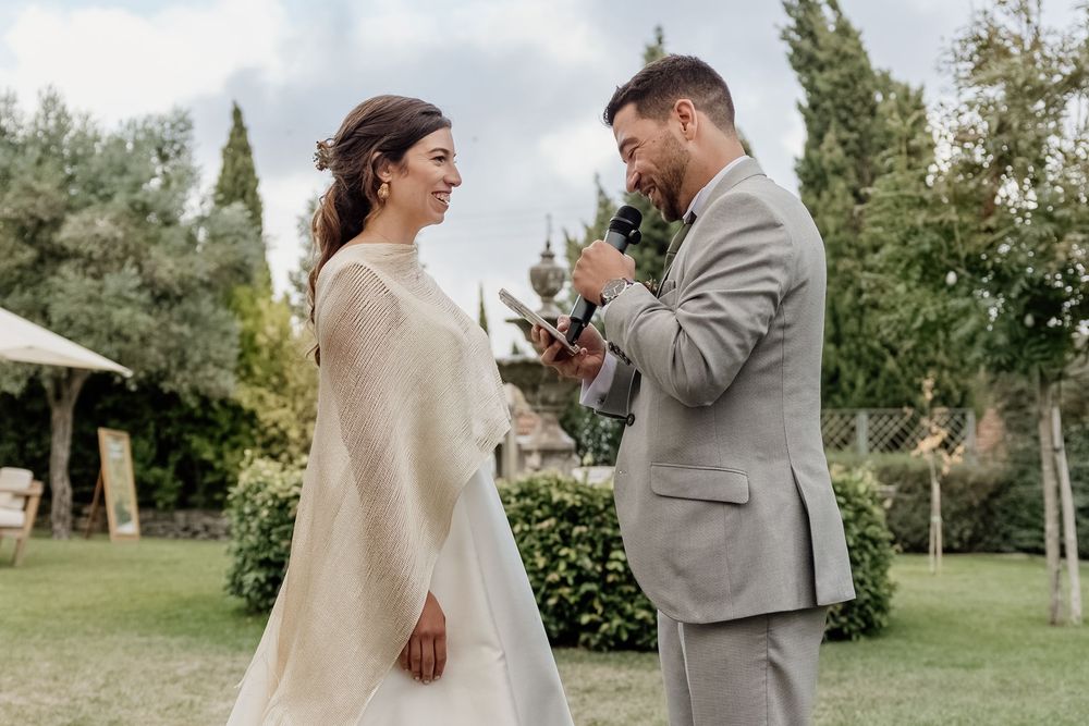 Groom in gray suit reads vows to bride in white dress and shawl during outdoor wedding ceremony.