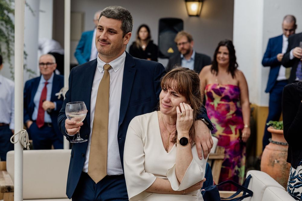 Emotional wedding guests in formal attire, woman wiping tears, man holding wine glass at elegant indoor ceremony.