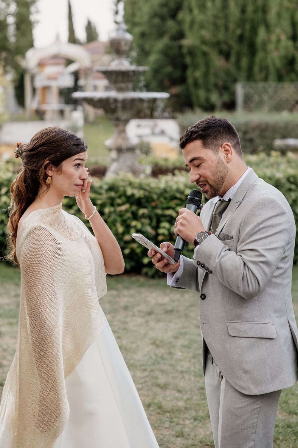 Groom reads wedding vows from phone while emotional bride wipes tears during outdoor garden ceremony.