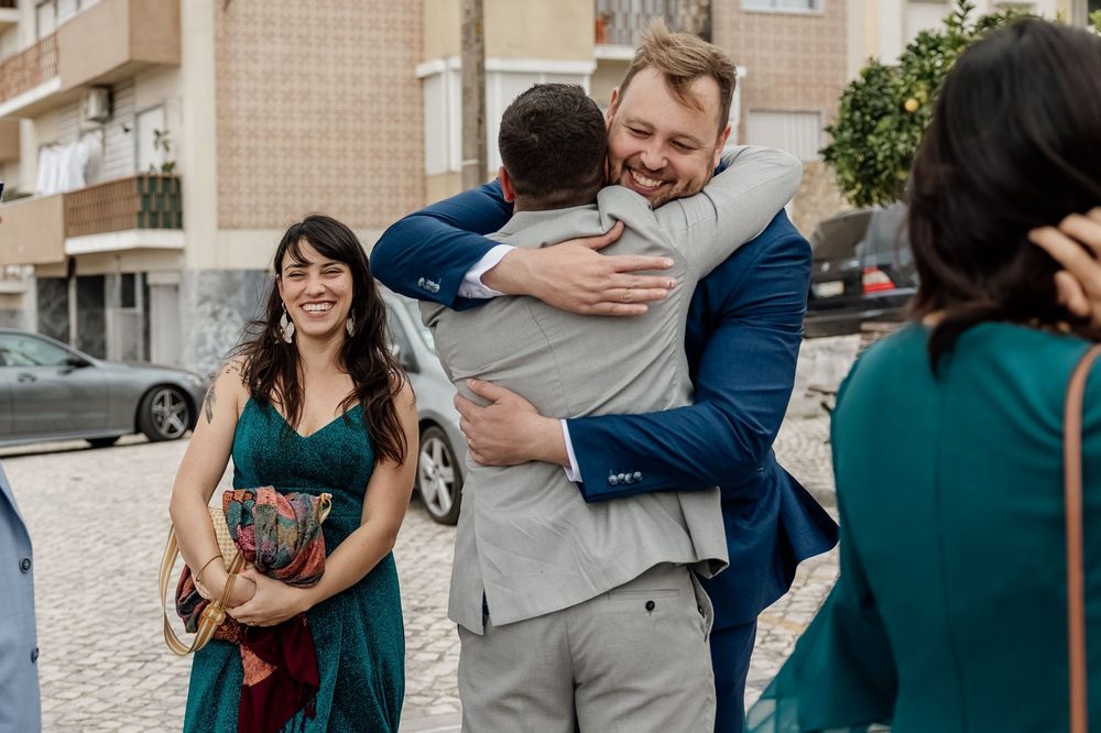 Two men share a warm hug at an outdoor wedding celebration while guests in elegant attire smile nearby.
