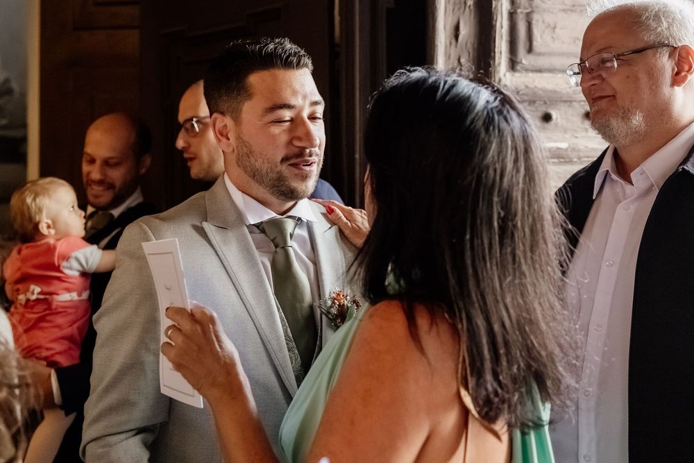 A groom in a gray suit smiles warmly while greeting guests at an elegant wedding reception indoors.