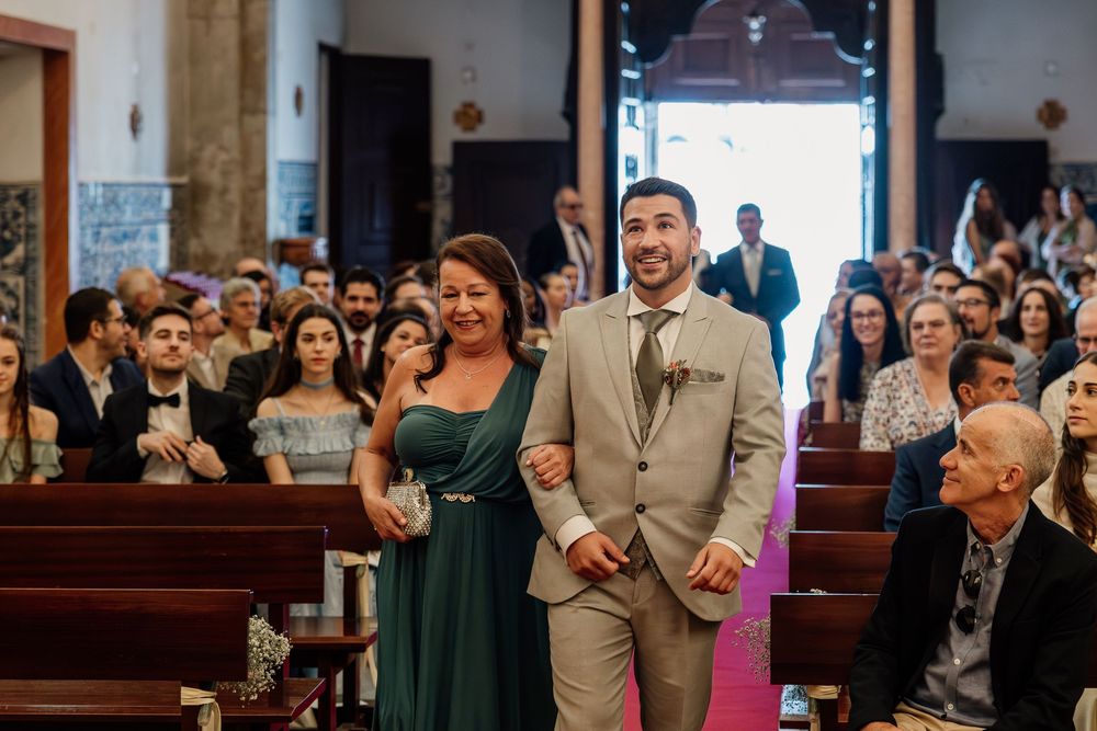 Groom in tan suit walks down church aisle with woman in green dress, guests seated in pews during wedding ceremony.