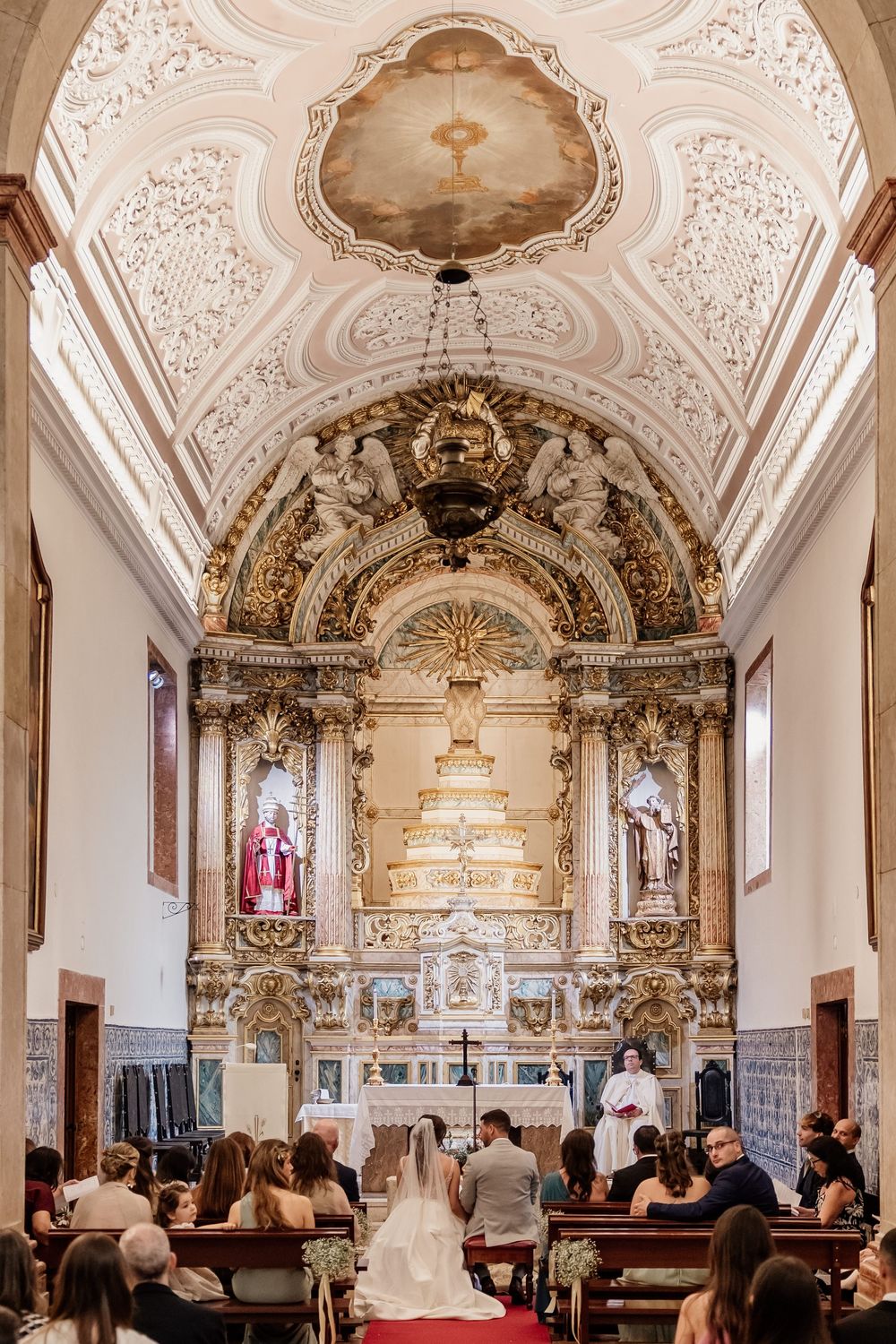 Elegant wedding ceremony in ornate Baroque church with gilded altar, bride and groom at altar, guests seated in pews.