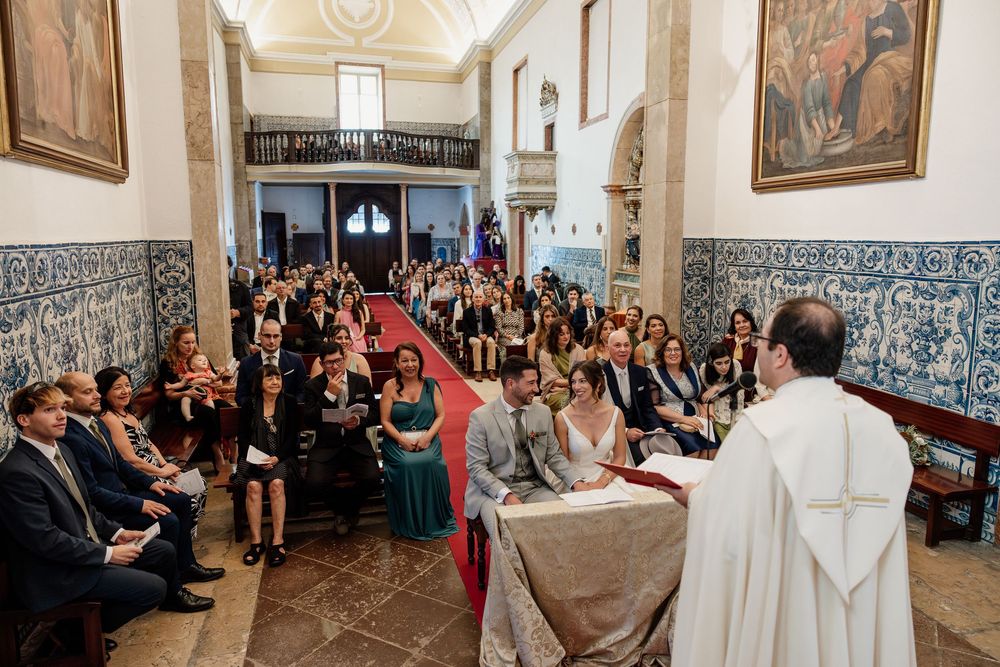 Wedding ceremony in ornate chapel with blue azulejo tiles, guests seated as couple exchanges vows before priest at altar.