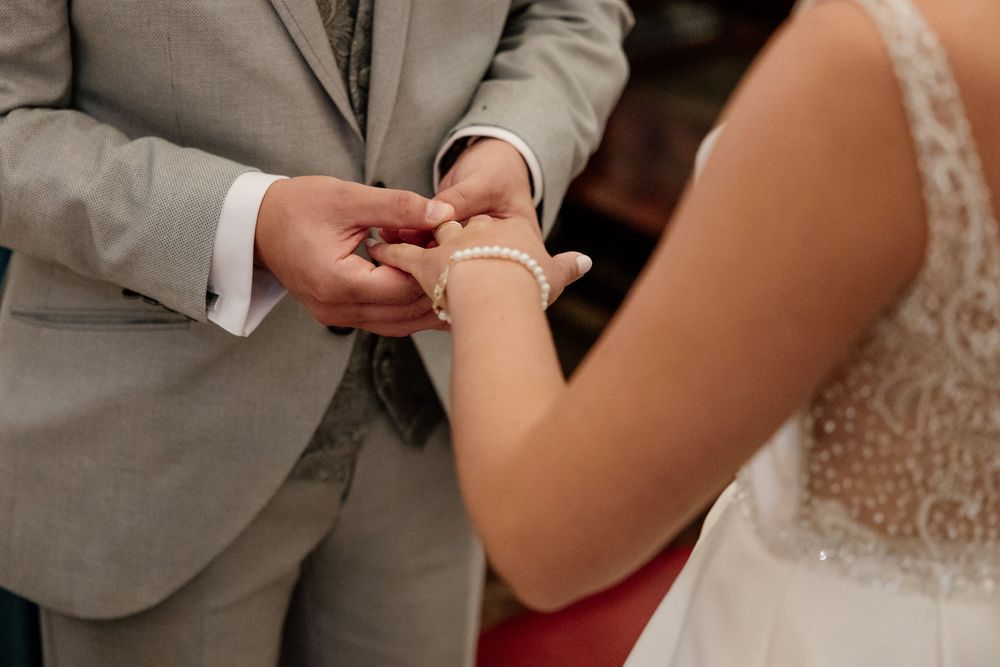 Groom in gray suit placing wedding ring on bride's hand with pearl bracelet during ceremony.