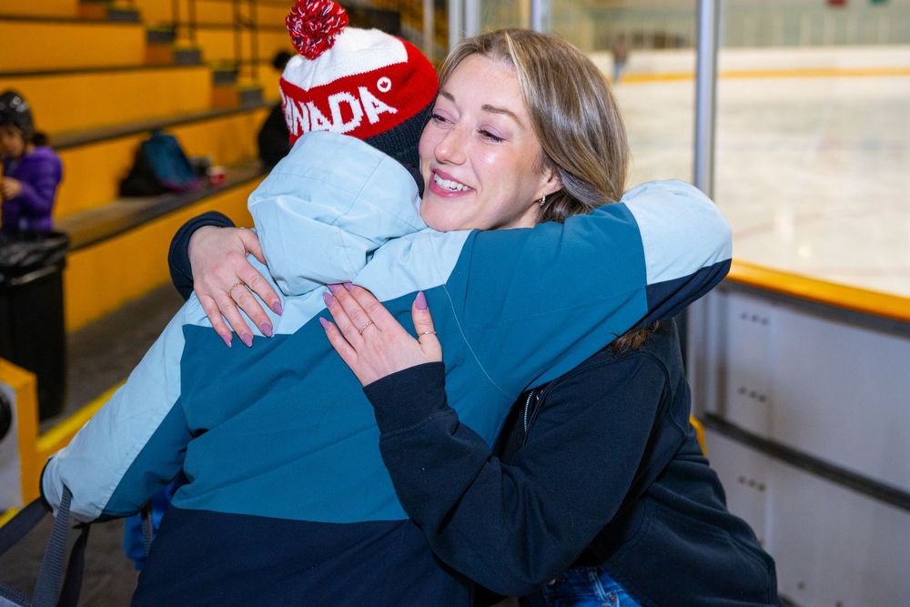 Two people share a warm embrace at an ice rink, with wooden bleachers visible in the background.