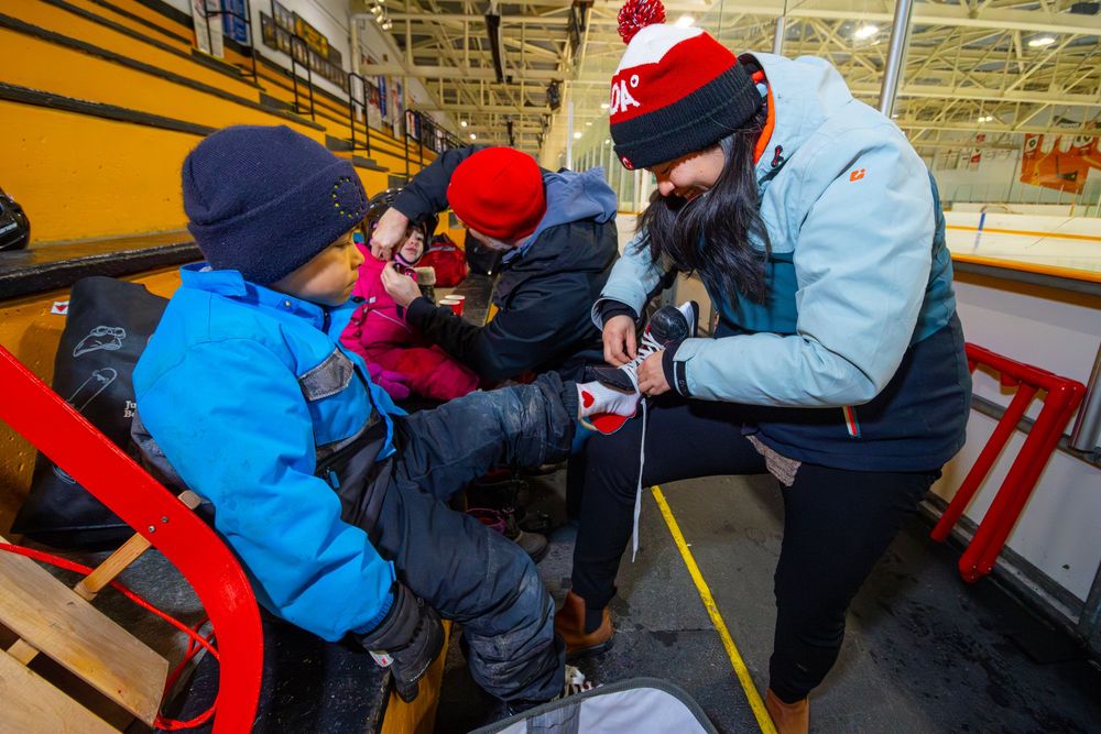 People in winter coats gather around an ice rink while someone helps another person lace up ice skates.