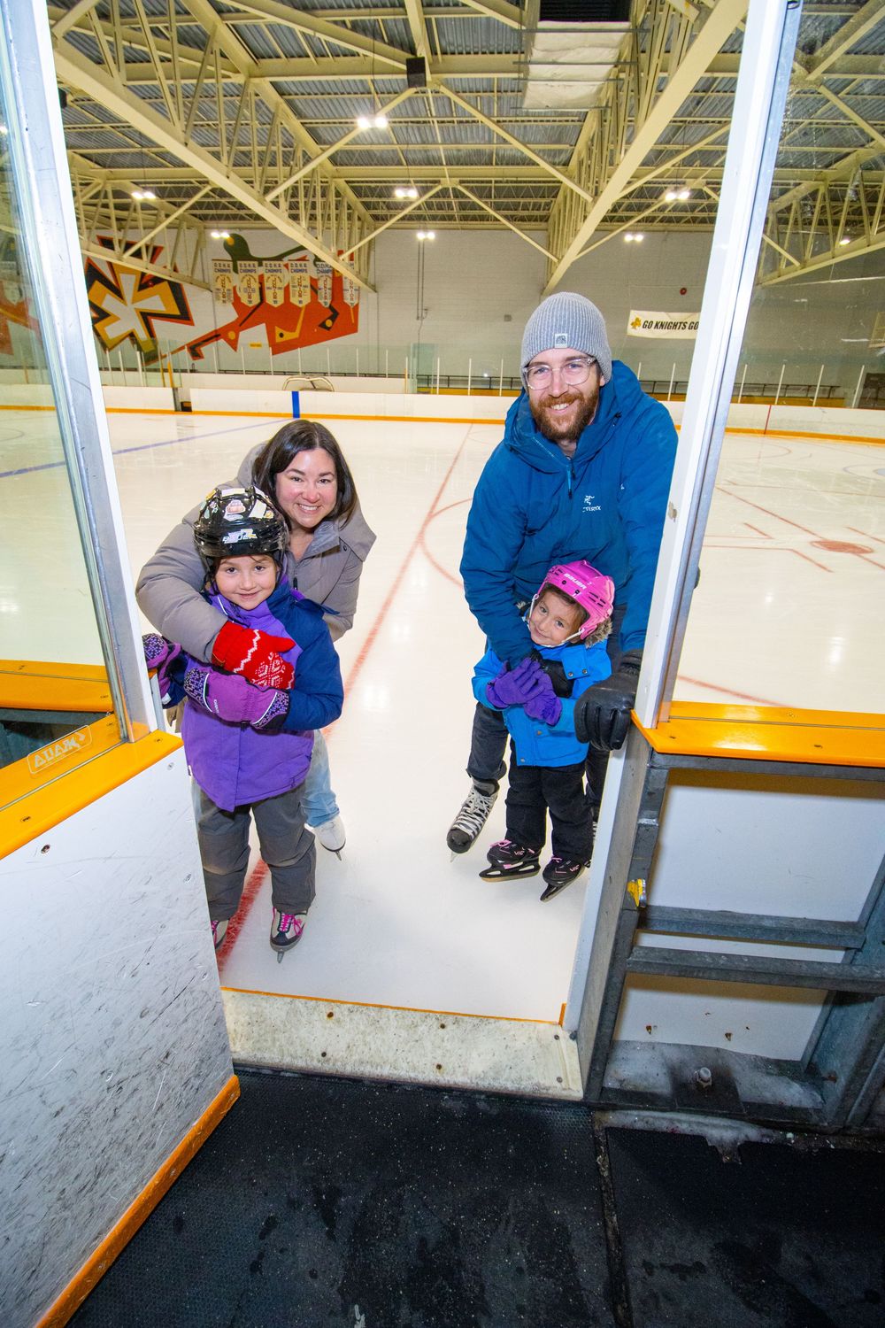 A group of skaters stand at the entrance to an indoor ice rink, bundled up in winter clothing.