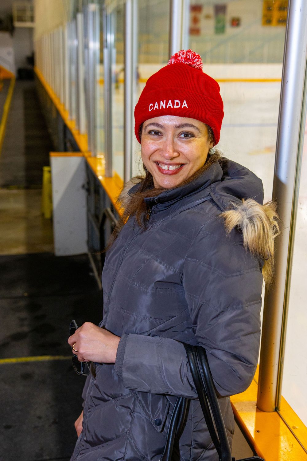 Person wearing a red Canada winter hat and grey winter coat smiles inside an ice rink facility.