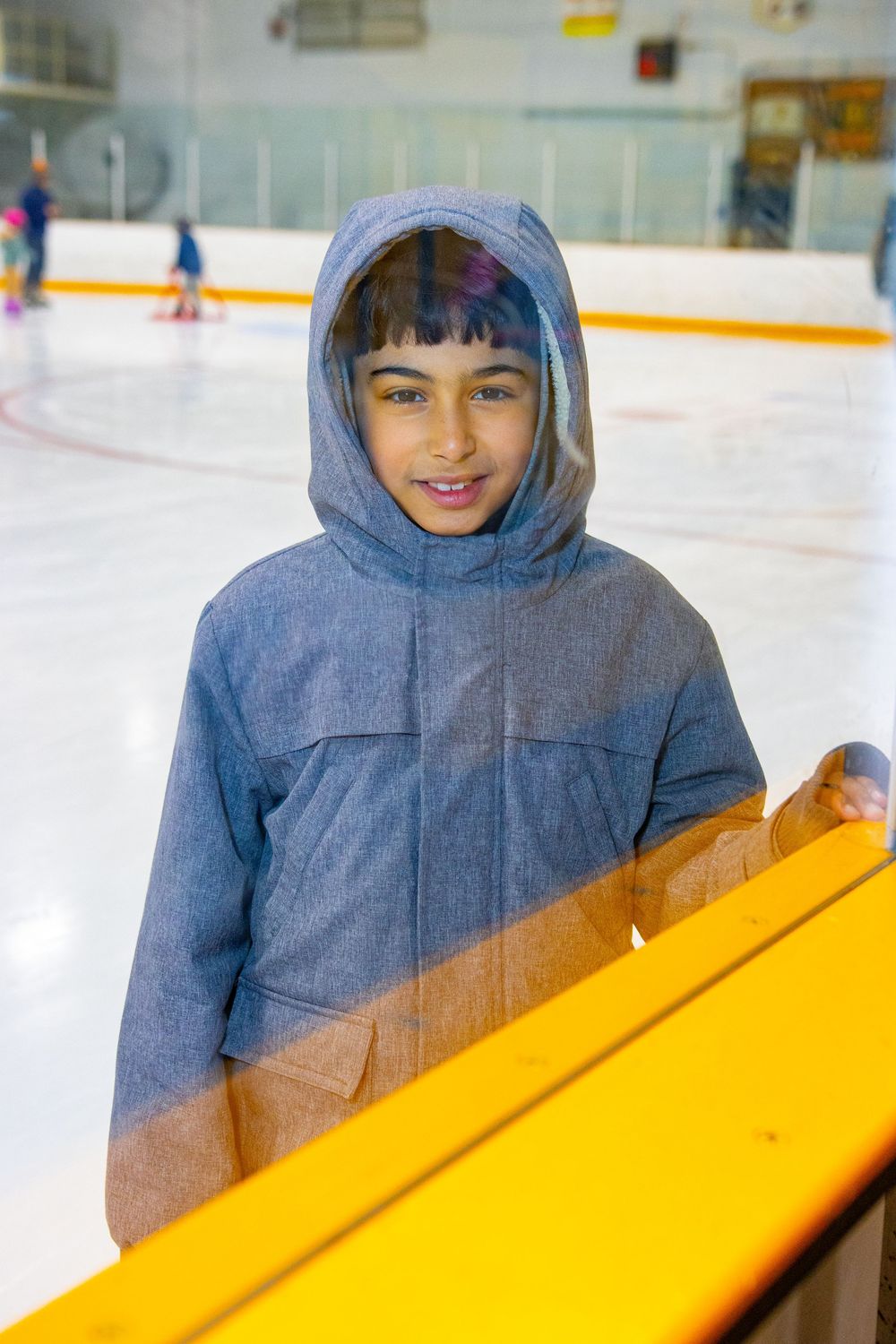 A person wearing a blue hooded sweatshirt leans on a yellow skating aid at an ice rink.
