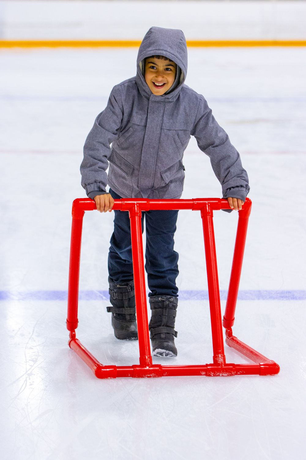 A child in a blue hoodie practices ice skating using a red training aid frame on an indoor rink.