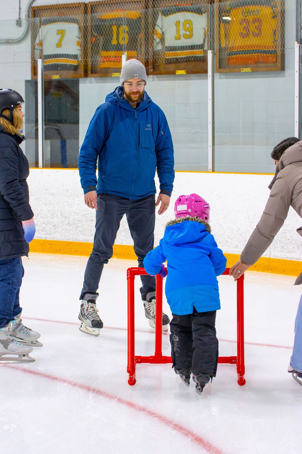 Group learning to ice skate with training aids at an indoor rink with hockey jerseys displayed on the wall.