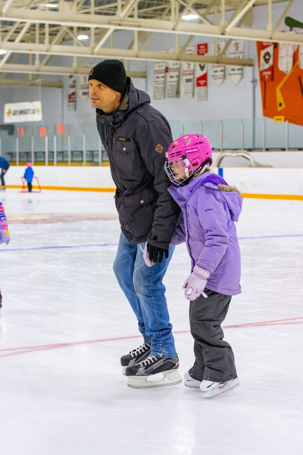 Adult and child practicing ice skating together at an indoor rink while holding hands for support and guidance.