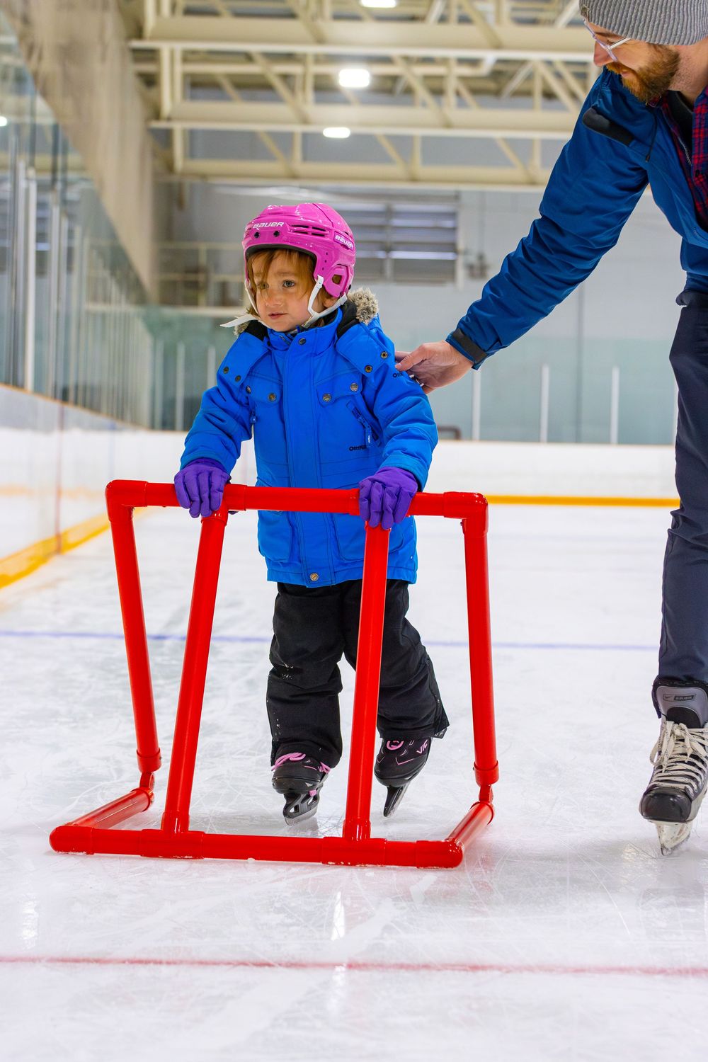 Young skater in pink helmet using red training aid while learning to ice skate.