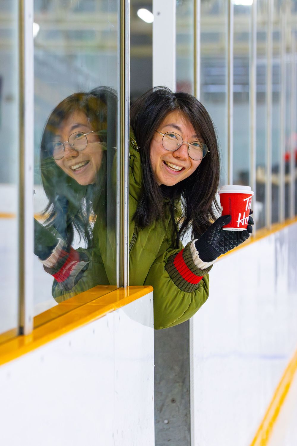 Person in green sweater peeks through hockey rink glass while holding red cup with reflection visible.