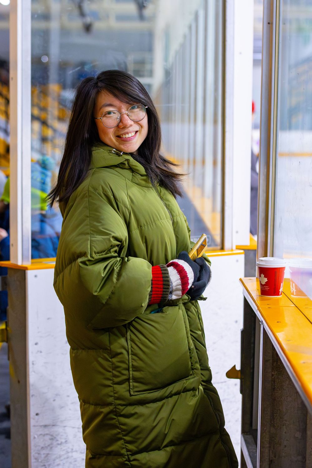 Person in a green winter coat standing by the glass barriers at an ice skating rink.