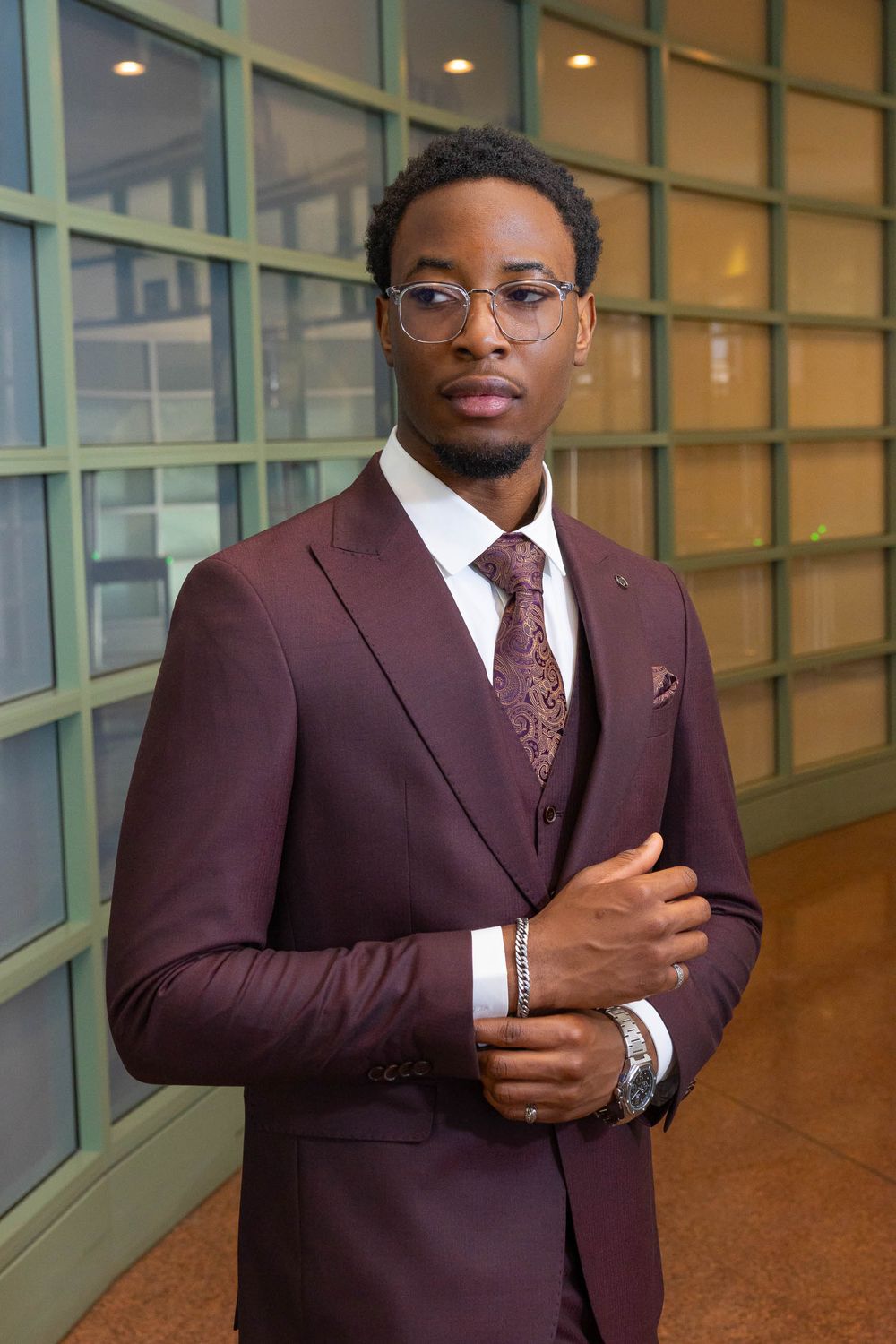 Professional in burgundy suit and glasses poses confidently in modern office hallway with glass walls.