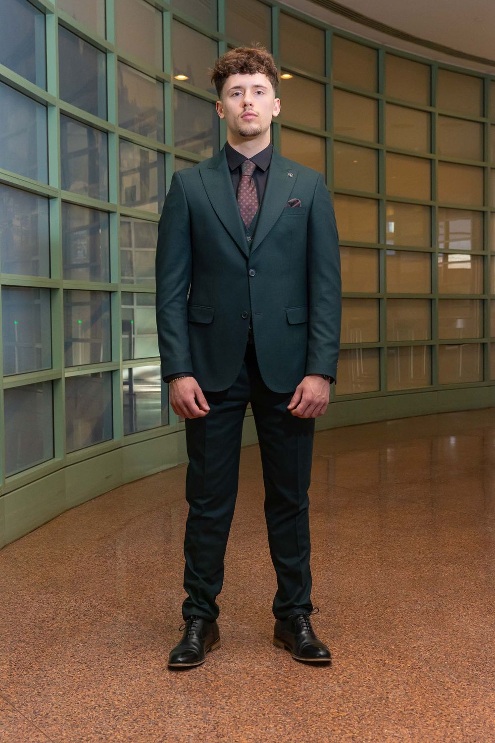 A person in a dark business suit stands in a modern office hallway with glass windows and brown flooring.