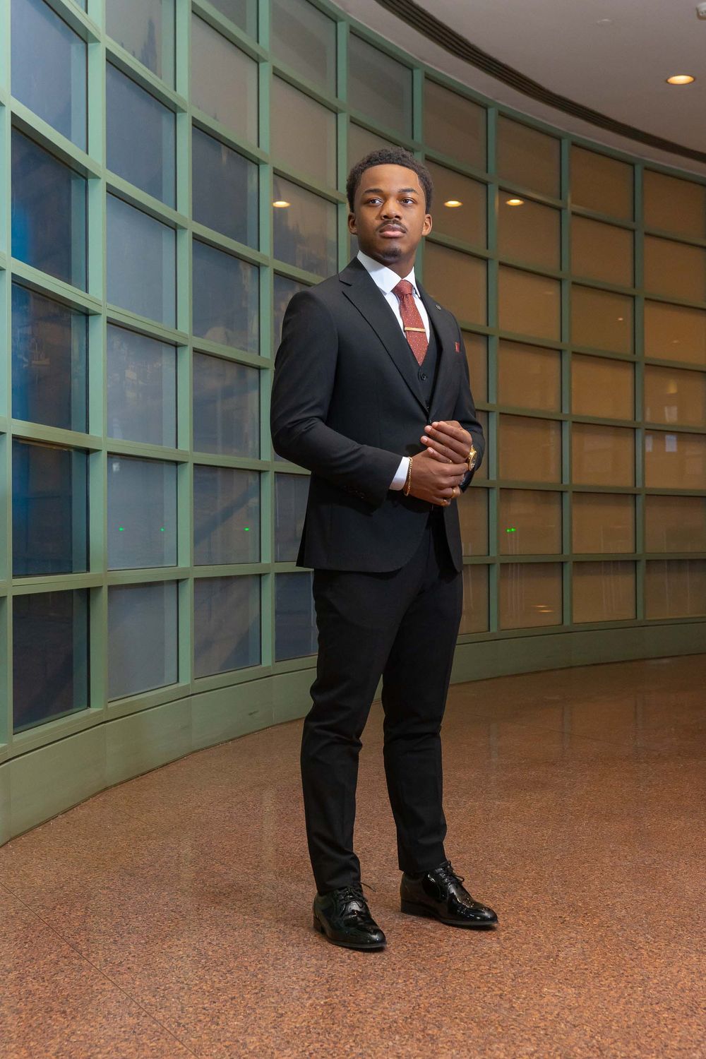 Well-dressed individual stands in an elegant curved corridor with floor-to-ceiling windows and warm lighting.