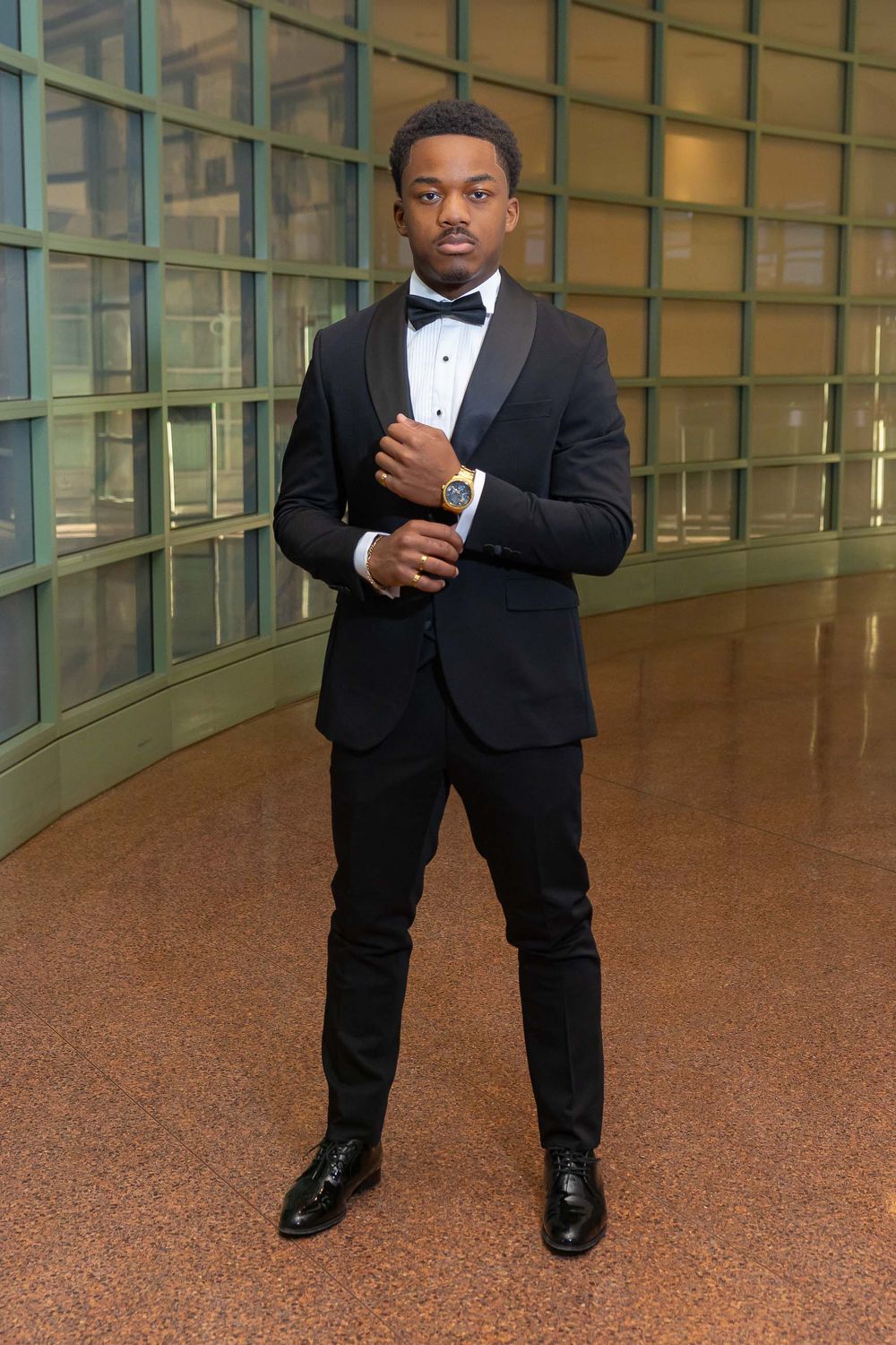 A person in a black tuxedo and bow tie poses confidently in an elegant hallway with tiled floors.