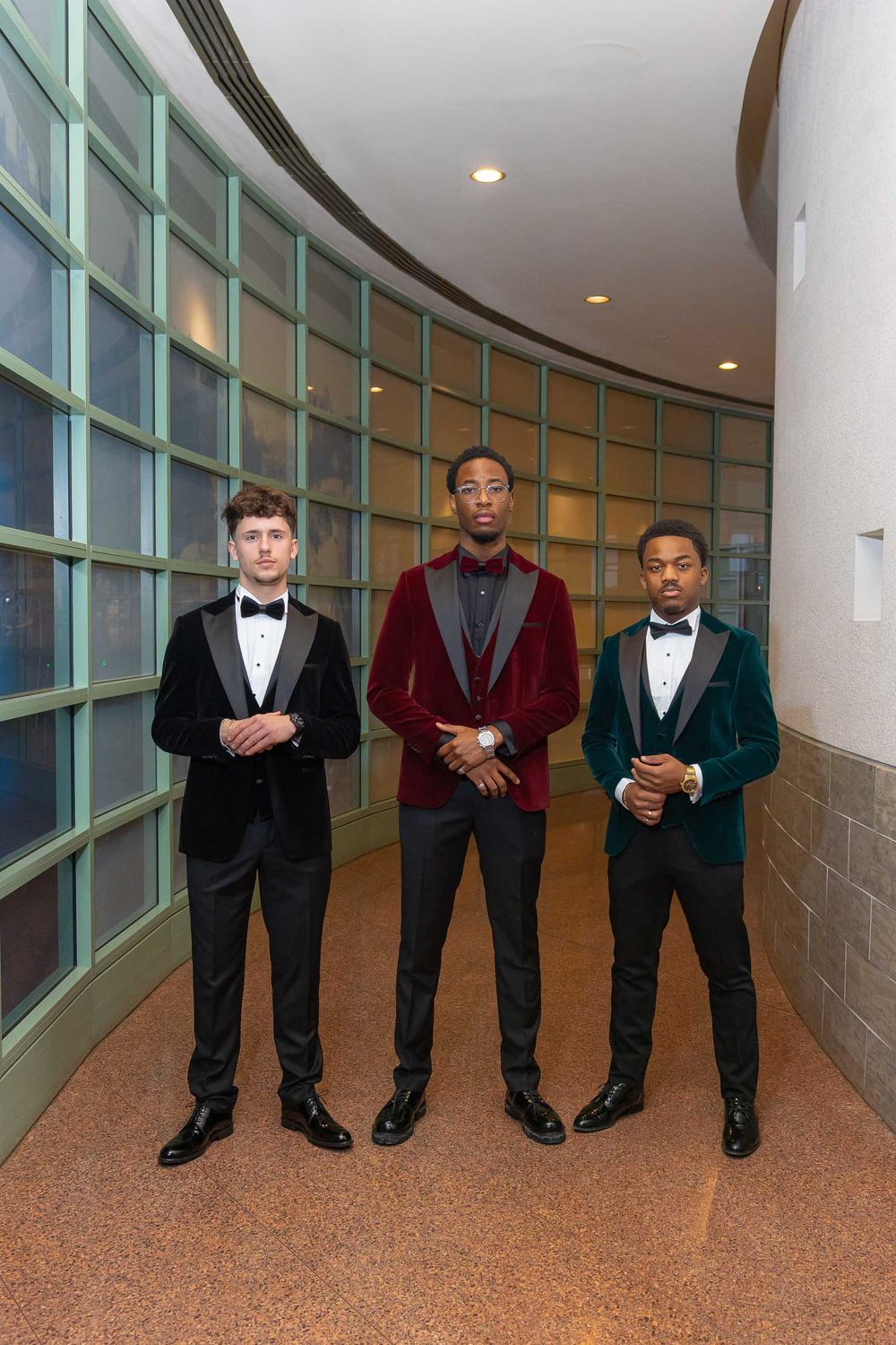 Three young men in formal attire stand in a curved hallway with glass block walls and recessed lighting.