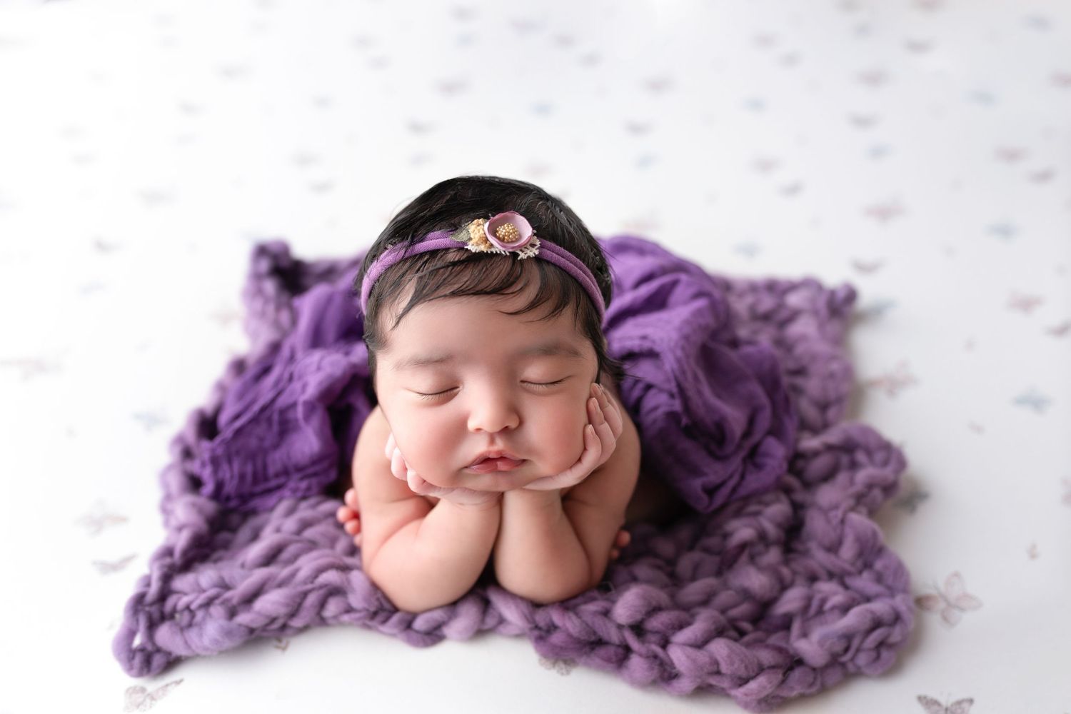 Sleeping newborn baby posed on purple knit blanket wearing delicate floral headband.