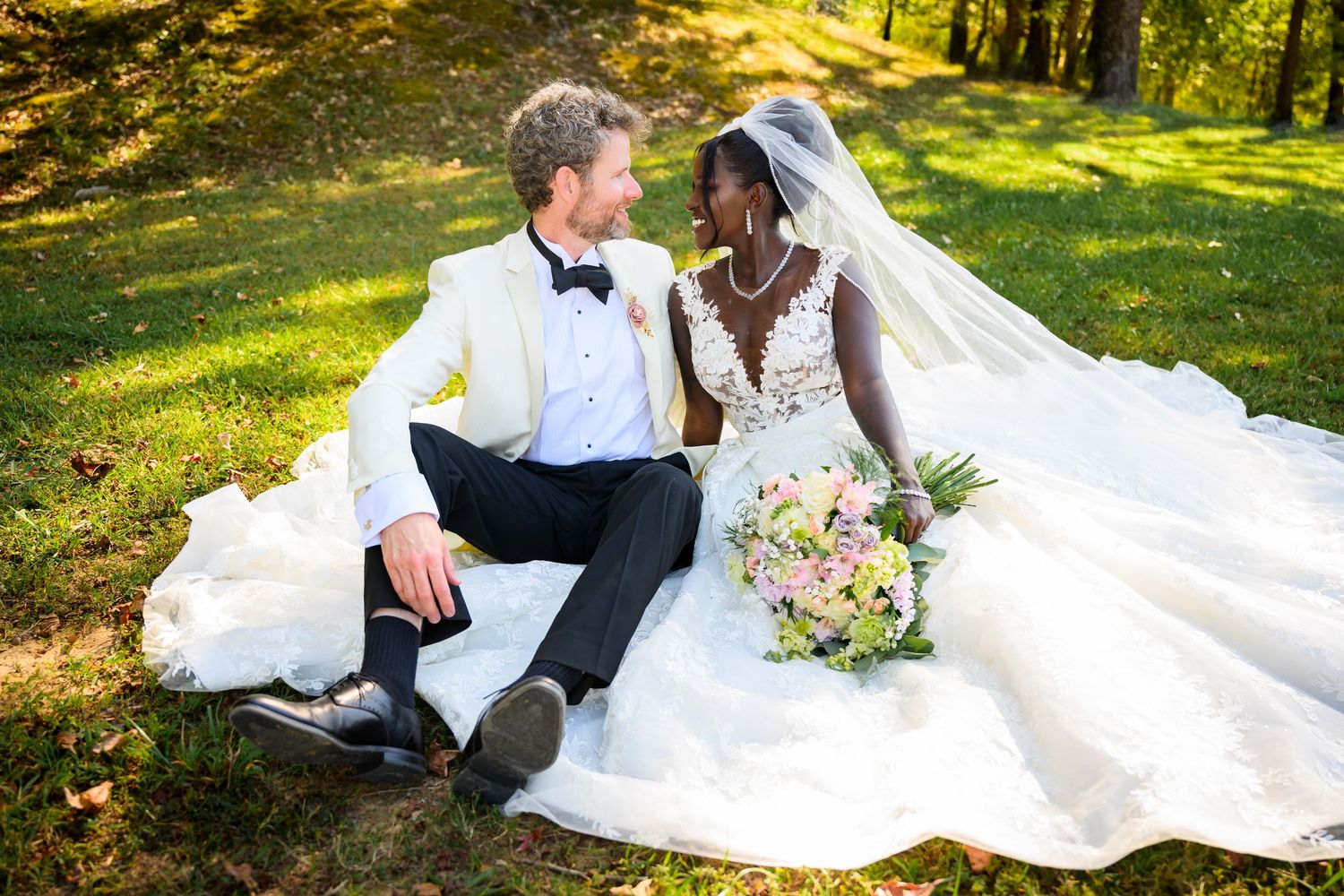 Interracial couple in wedding attire sitting on grass, bride in white gown with veil holding bouquet, groom in white tuxedo.