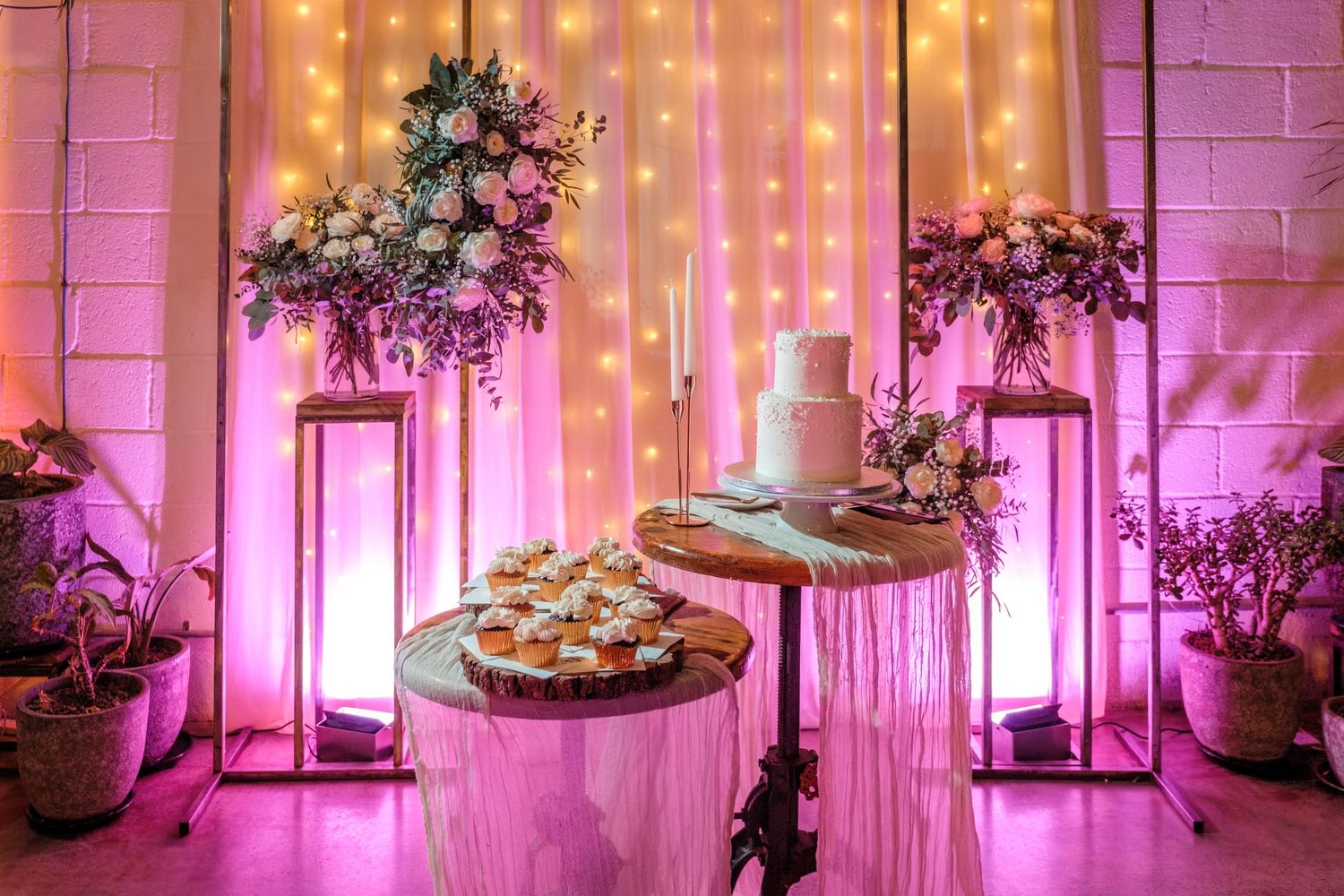 Elegant wedding dessert table with tiered cake, cupcakes, floral arrangements, and warm fairy lights backdrop.