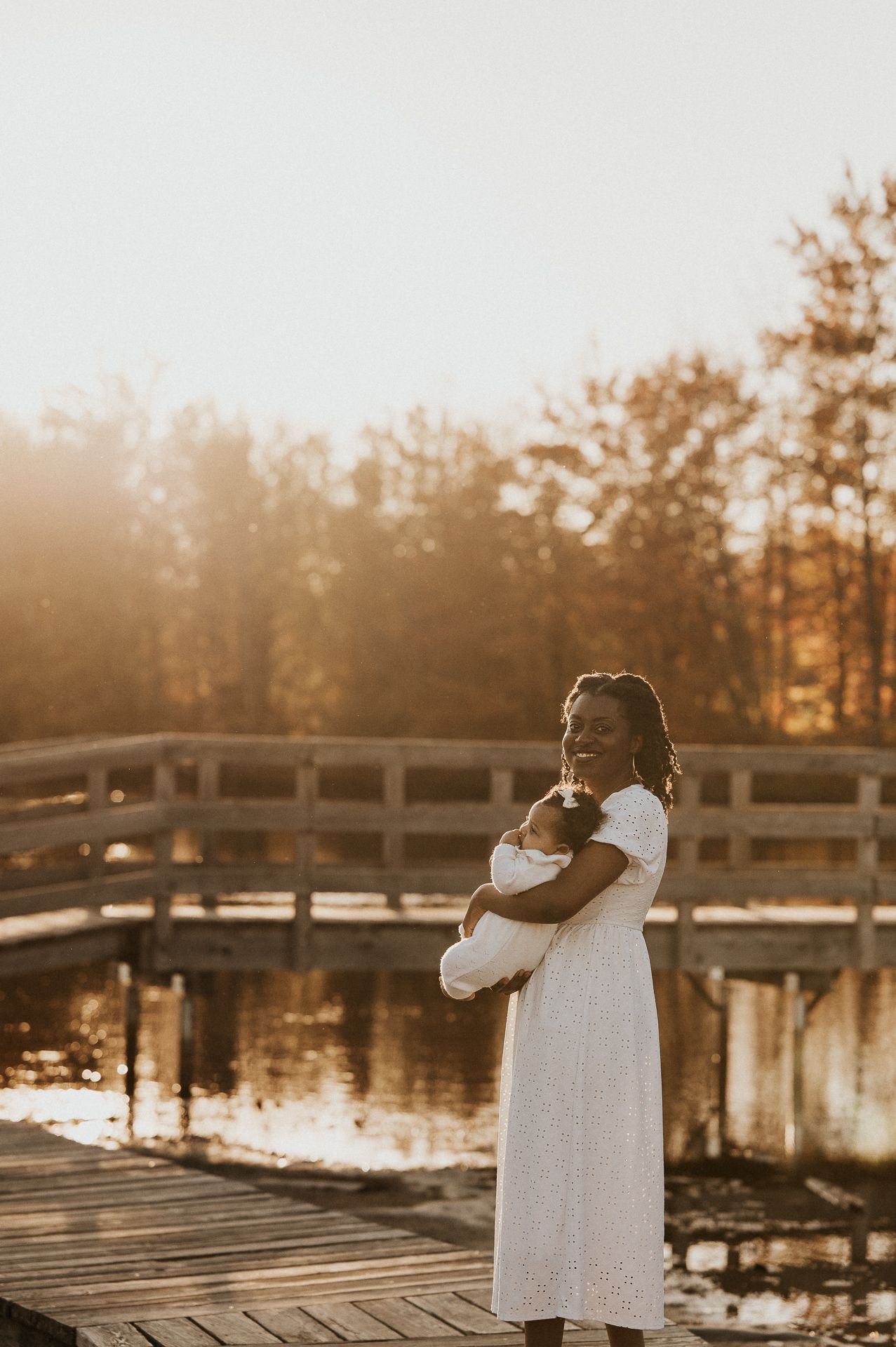 Maman et bébé près d’un plan d’eau au coucher du soleil, arbres en arrière-plan. Séance famille Rive-Sud de Montréal.