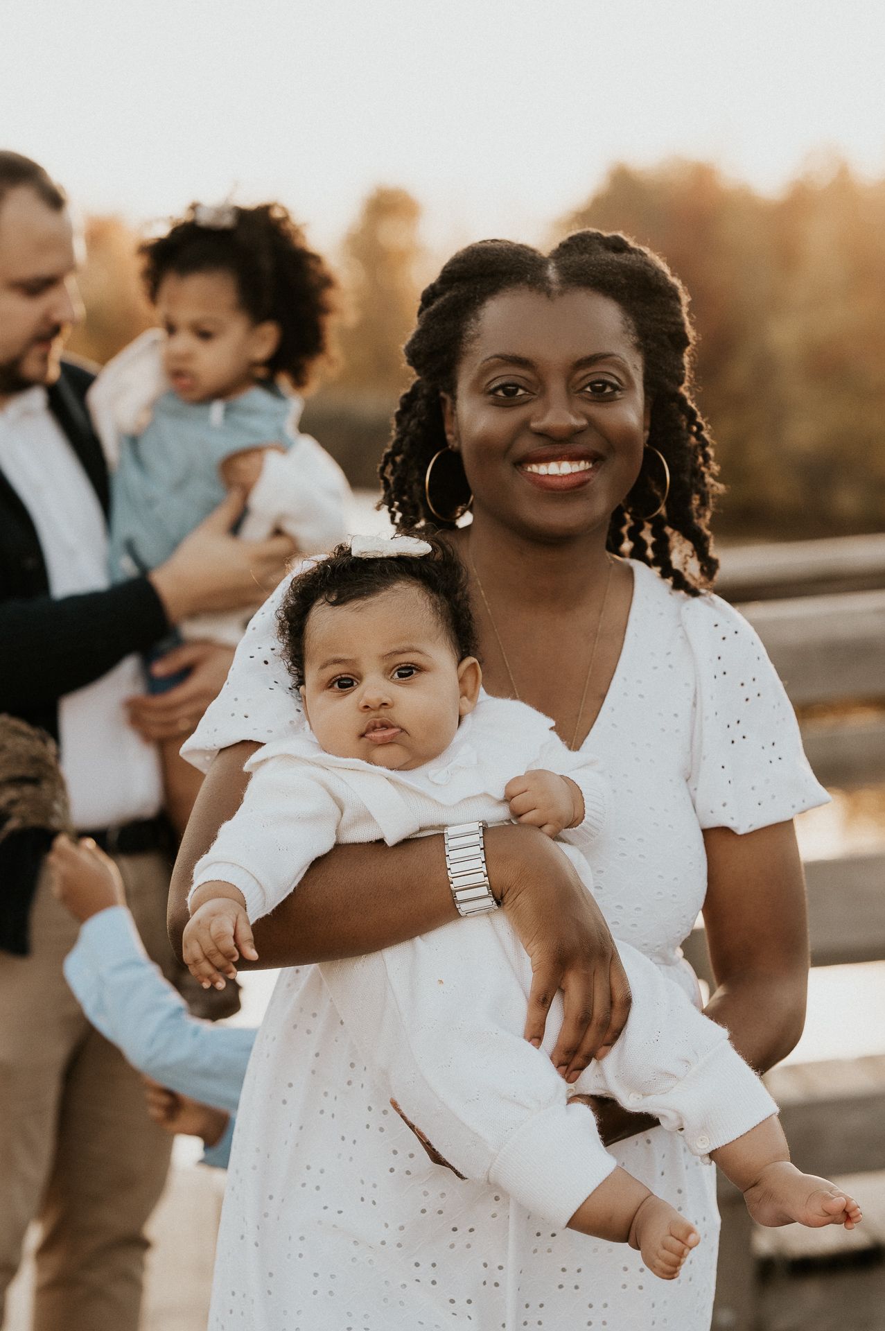 Maman tenant son bébé en extérieur, avec la famille en arrière-plan.