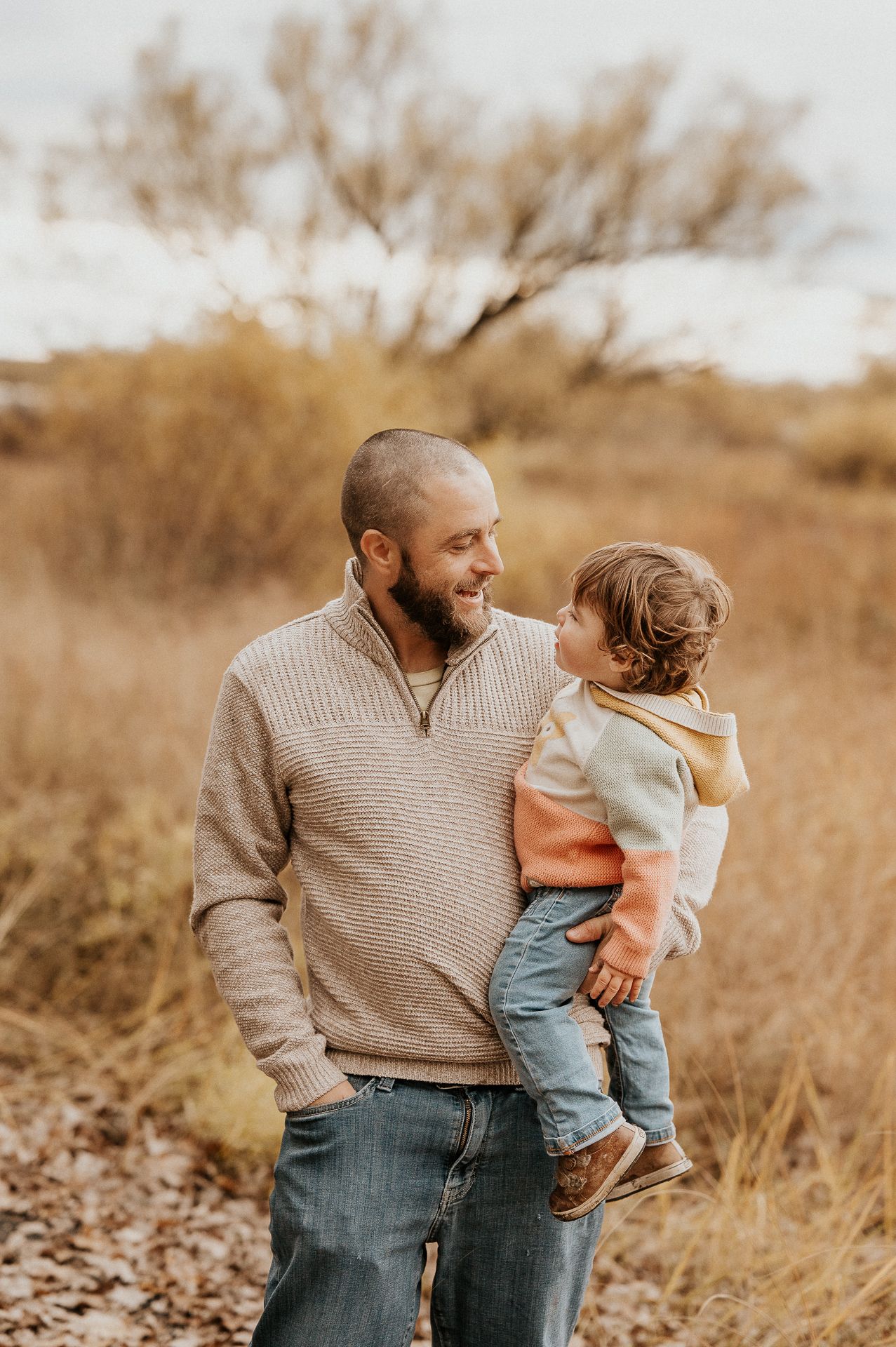 Adulte tenant un enfant dans un champ aux tons beige, lumière naturelle douce. Séance famille sur la Rive-Sud de Montréal.