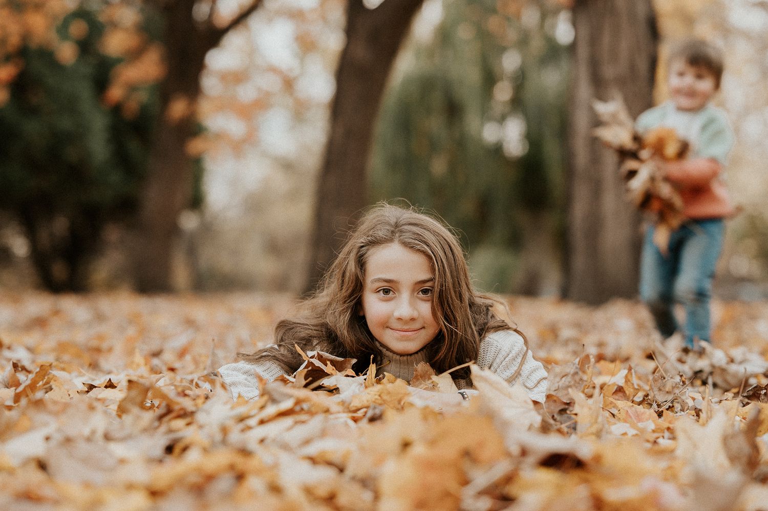Enfant couché dans des feuilles d’automne en forêt, arbres flous en arrière-plan.