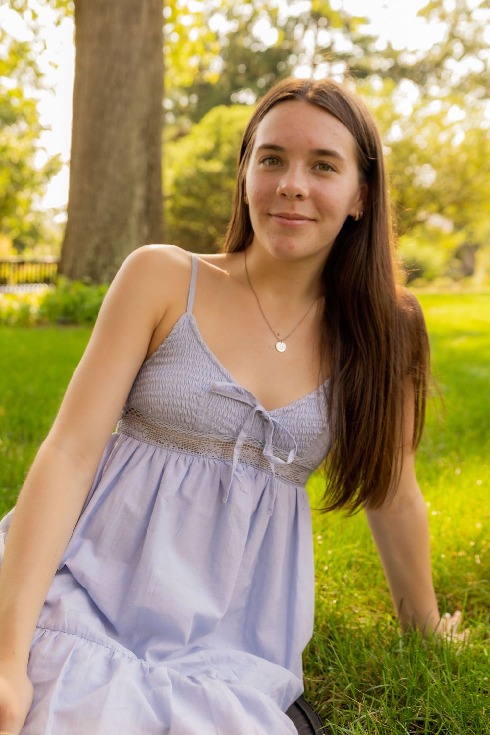 A person in a light purple sundress sits on grass in a sunny garden setting with trees in the background.