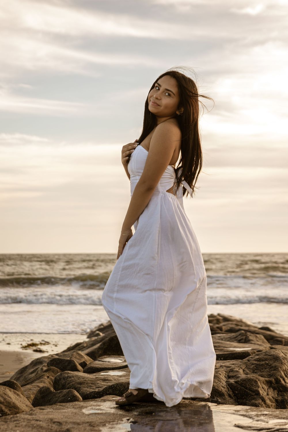A person in a flowing white dress poses gracefully on coastal rocks at sunset by the ocean.