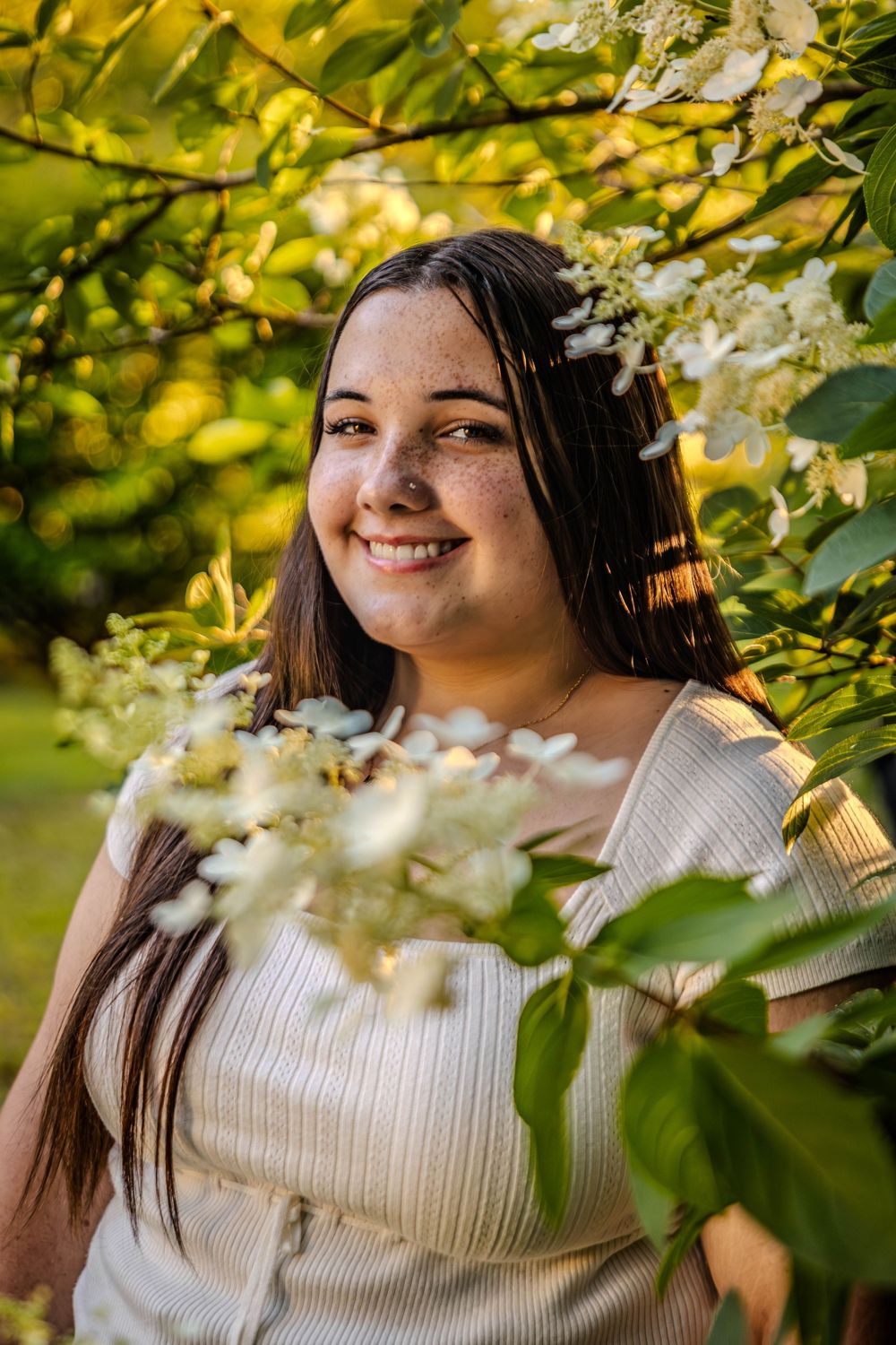 A portrait in a white dress surrounded by spring blossoms and green foliage in warm sunlight.