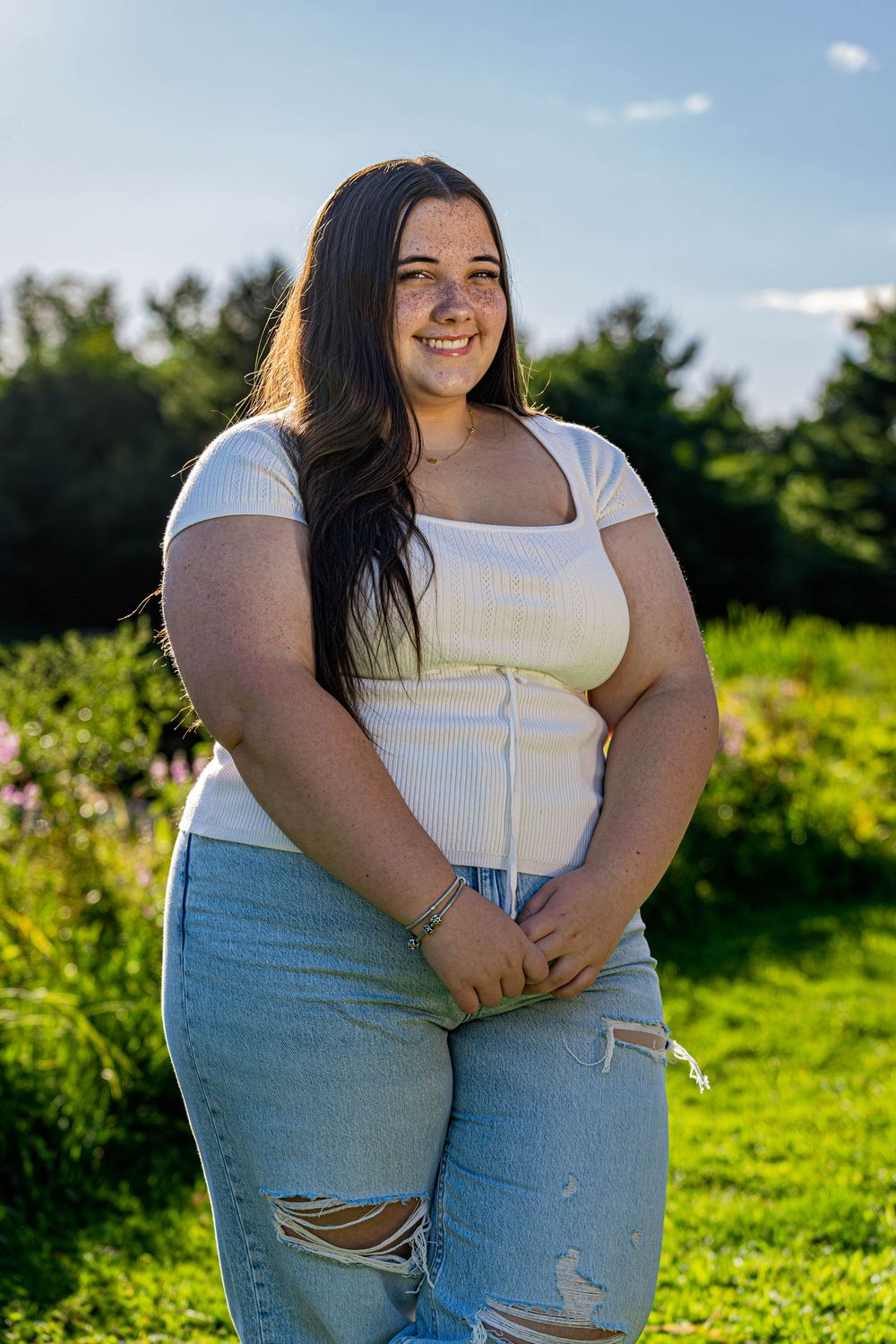 A person in a white top and ripped jeans poses outdoors in a sunny field with green foliage in the background.