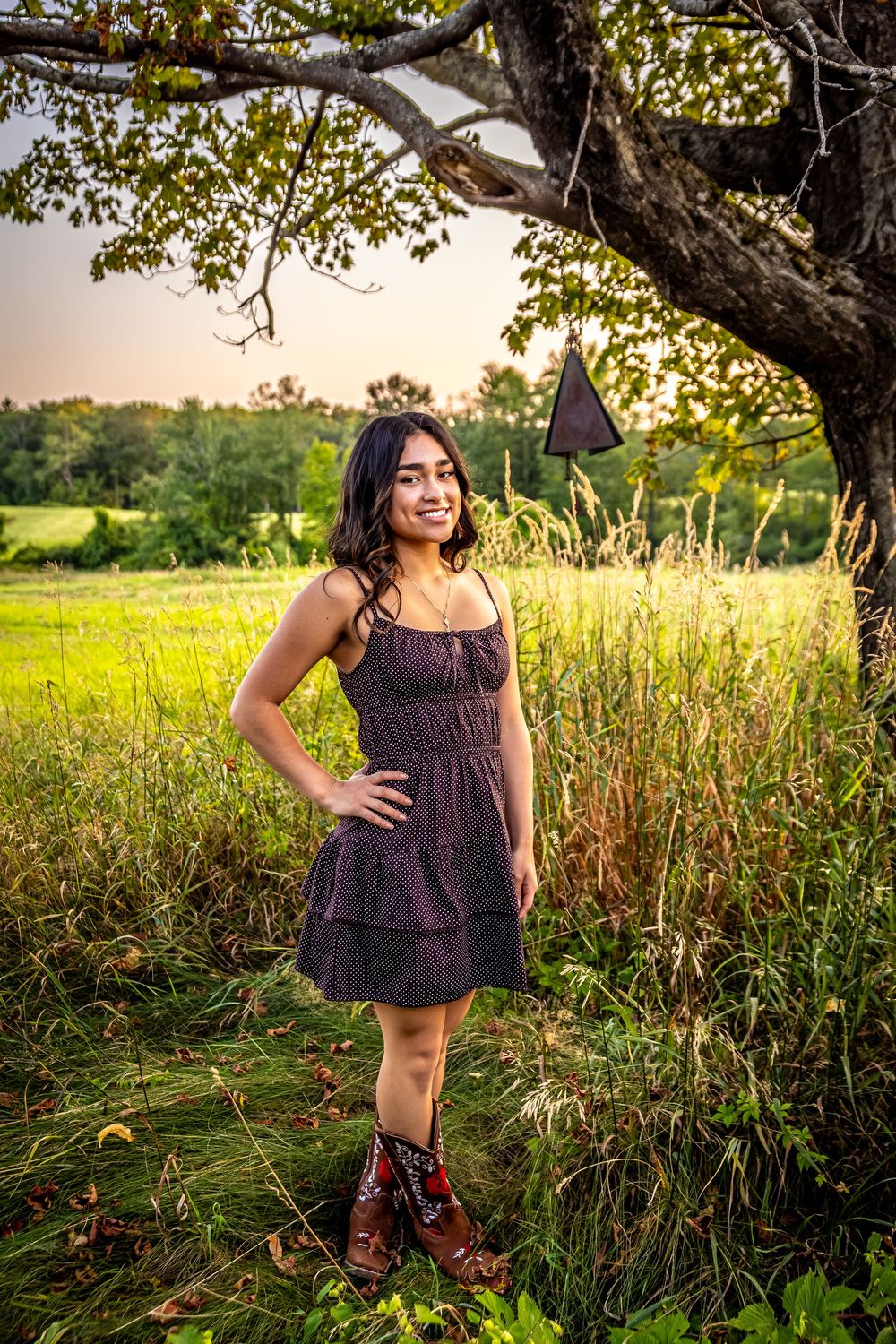 A person in a purple dress and boots poses in a sunlit meadow during golden hour with tall grass and trees.