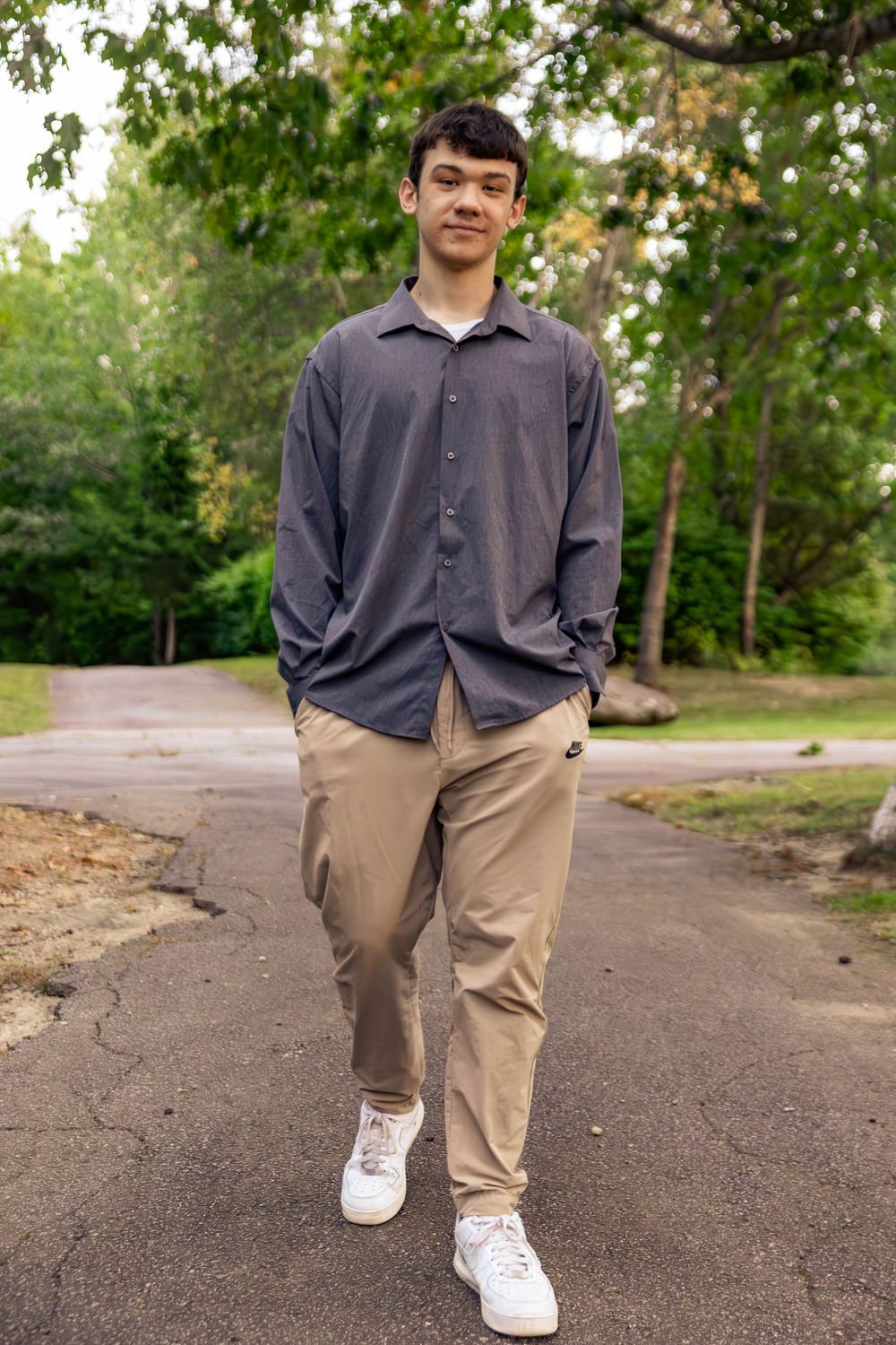 A person in a grey button-down shirt and khaki pants stands on a paved path surrounded by green trees.