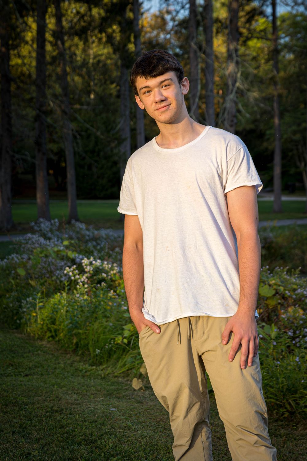 Person in white t-shirt and khaki pants poses casually in outdoor garden setting during golden hour.