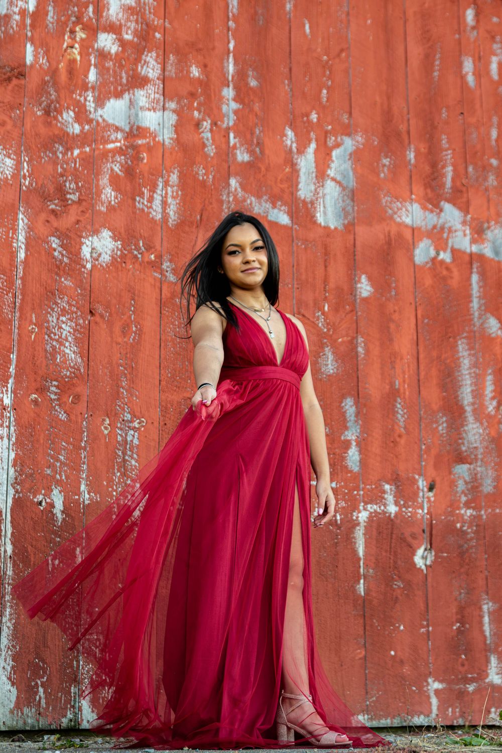A flowing burgundy tulle gown with deep v-neckline and side slit stands against a weathered orange wall.
