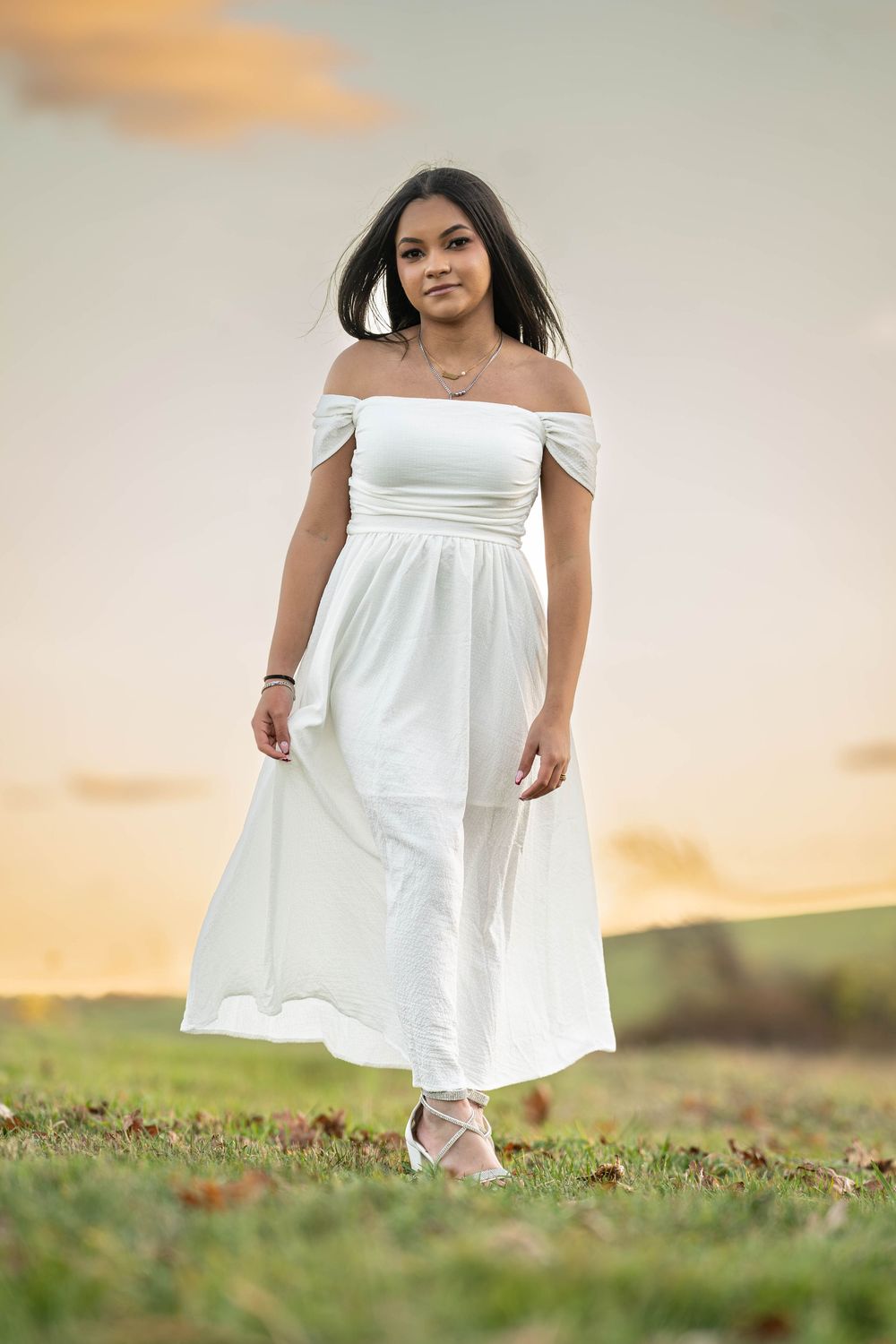 A person in a flowing white off-shoulder dress stands on a grassy hill during golden hour sunset.