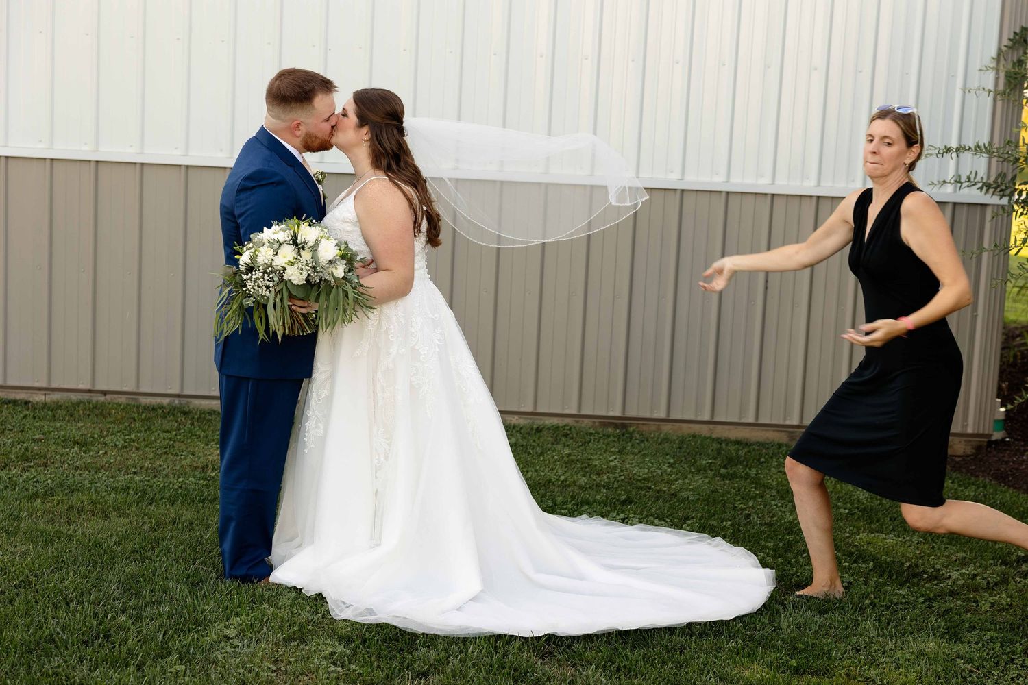 A couple in wedding attire share a romantic moment while someone in a black dress playfully poses in the background.