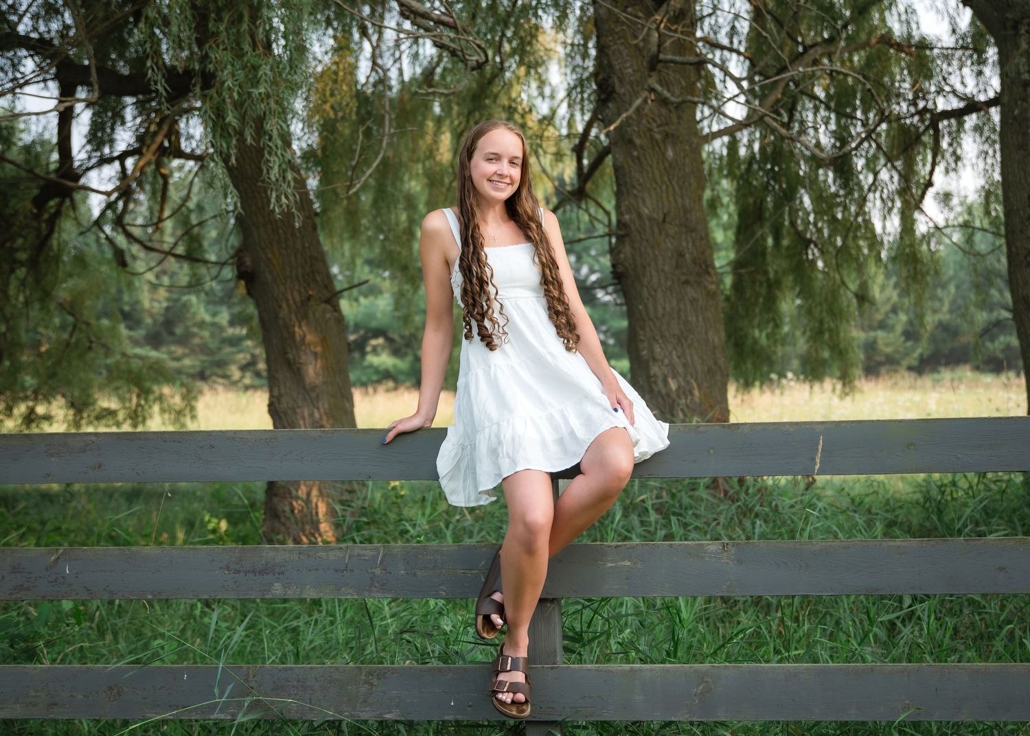 A woman in a white dress stands near a wooden fence in a scenic outdoor setting with willow trees.
