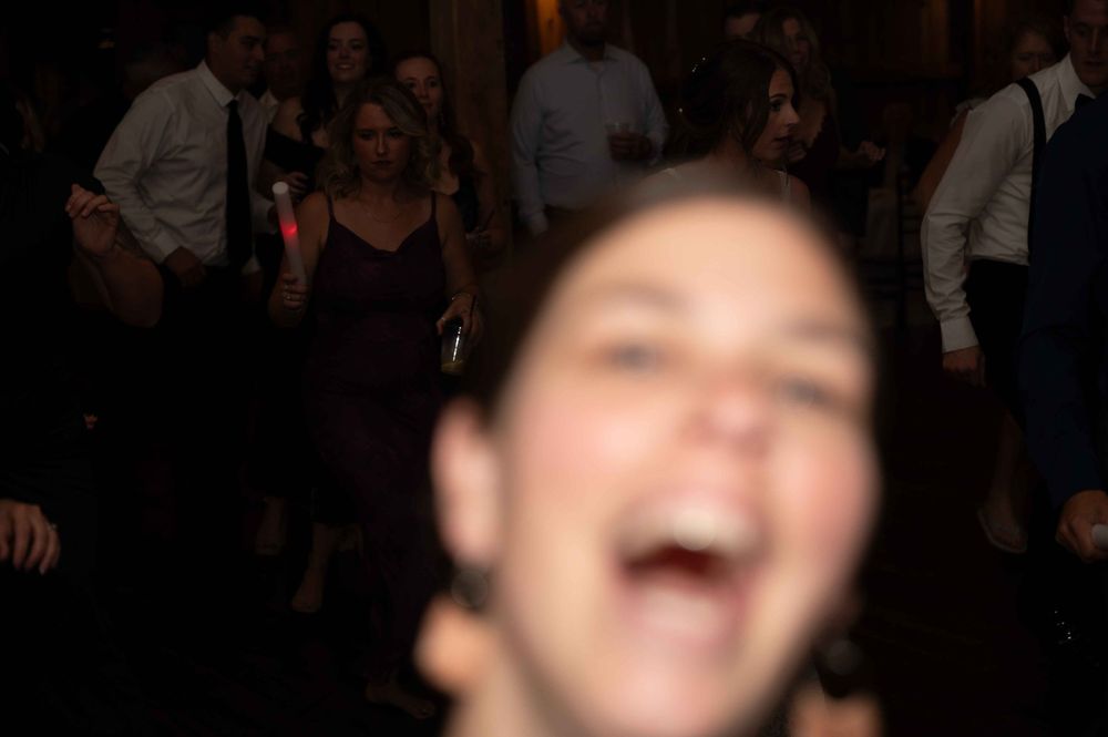 A person laughs joyfully while sitting in a dimly lit room with others visible in the background.